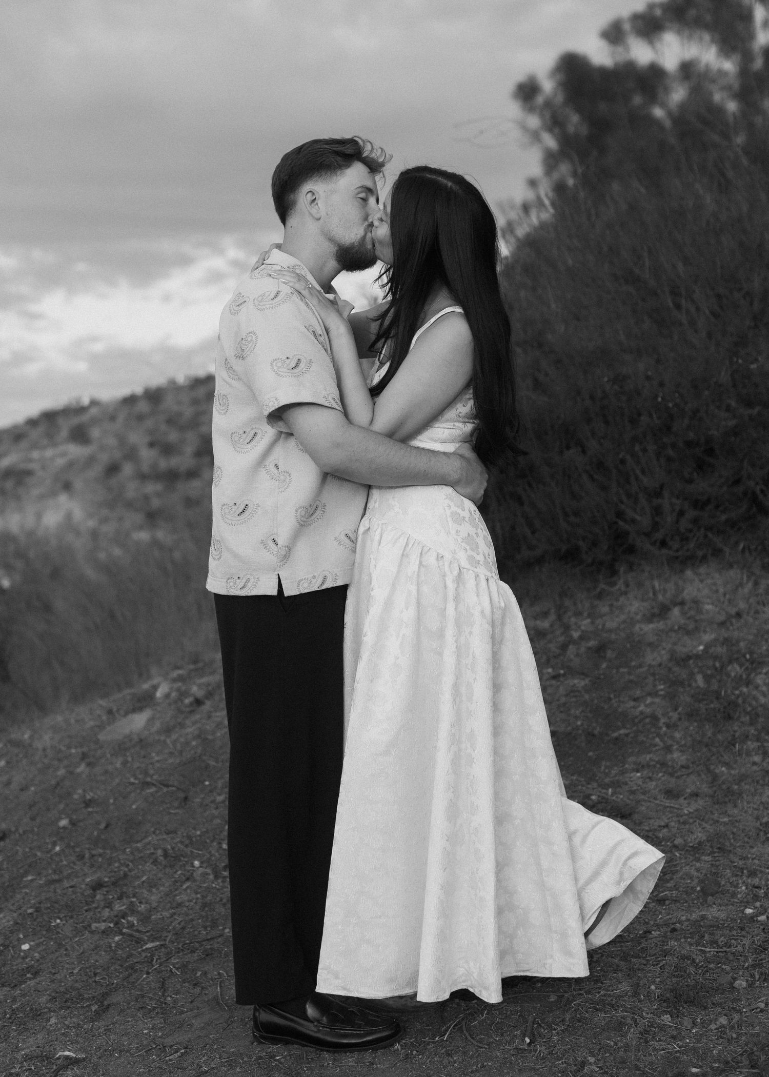 Black and white portrait of a couple sharing tender moments on a hillside.