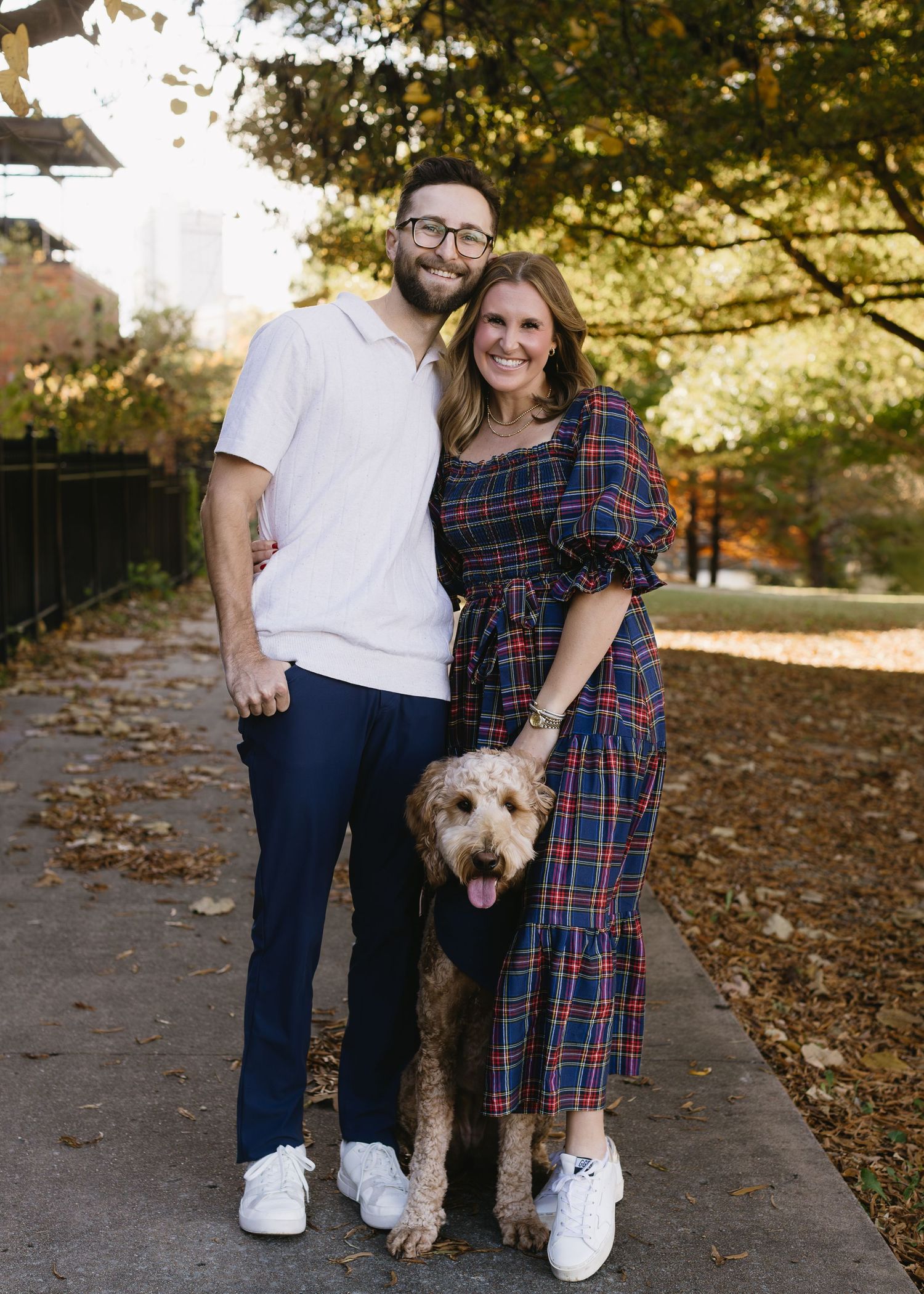 A couple poses with their fluffy goldendoodle dog on a leaf-covered path during an autumn photoshoot.