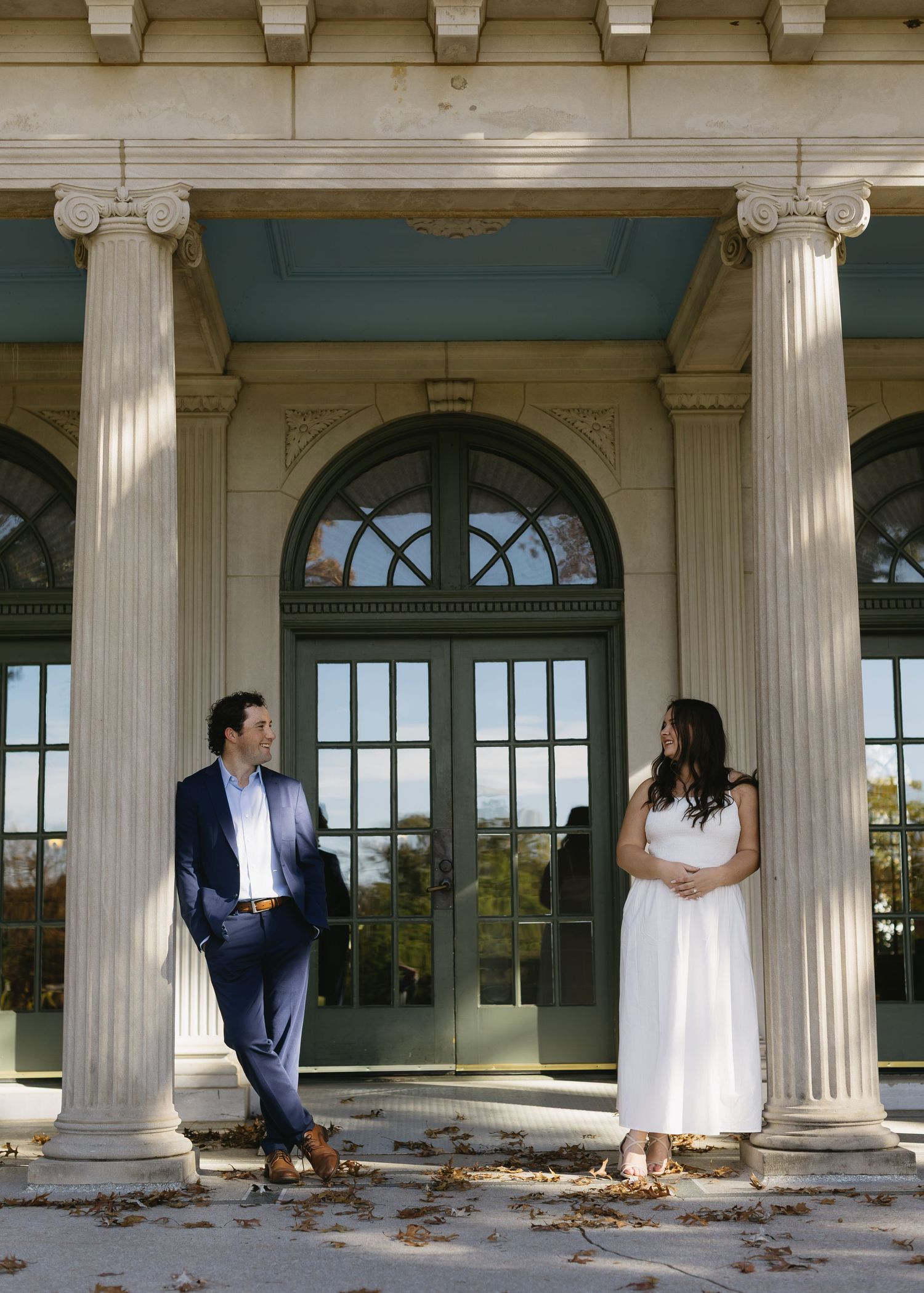 A couple in formal wear stands between elegant white columns of a historic building.