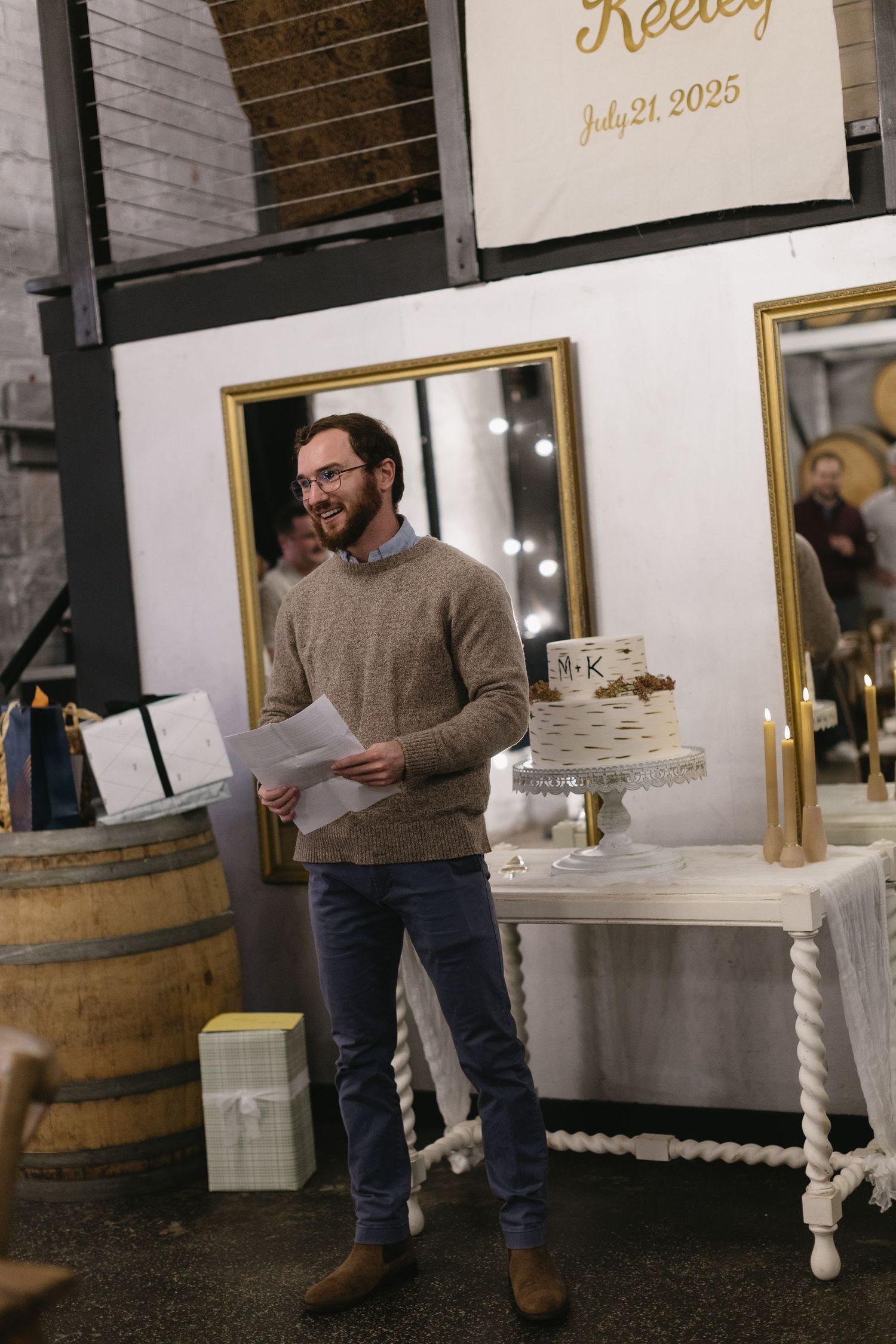 Speaker delivering remarks at wedding reception with wine barrel and dessert table in background.