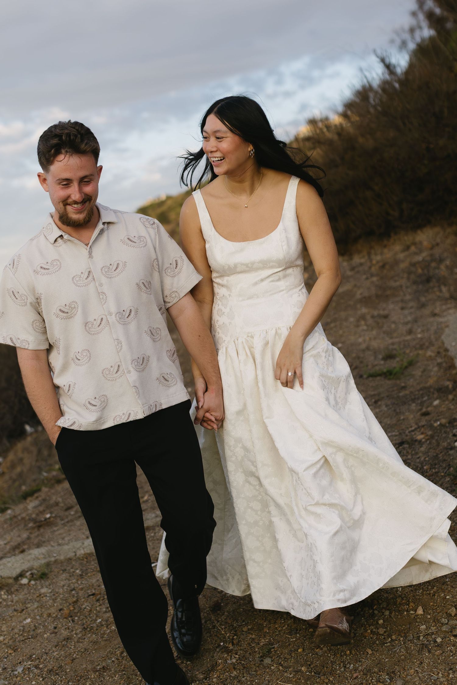 Couple walks hand in hand along a dirt path wearing coordinated white outfits for their photoshoot.