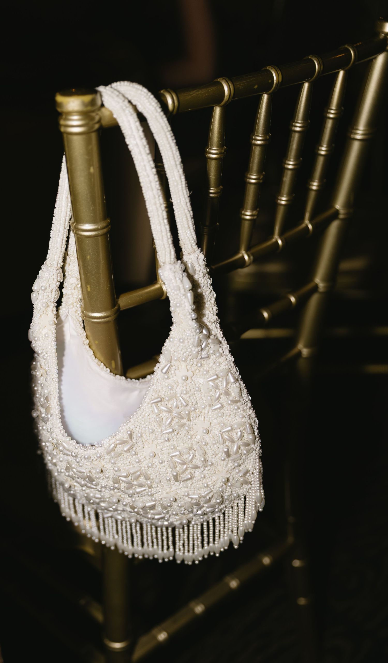 Delicate white lace handbag with beaded fringe detail hanging on gold metal railing against dark background.