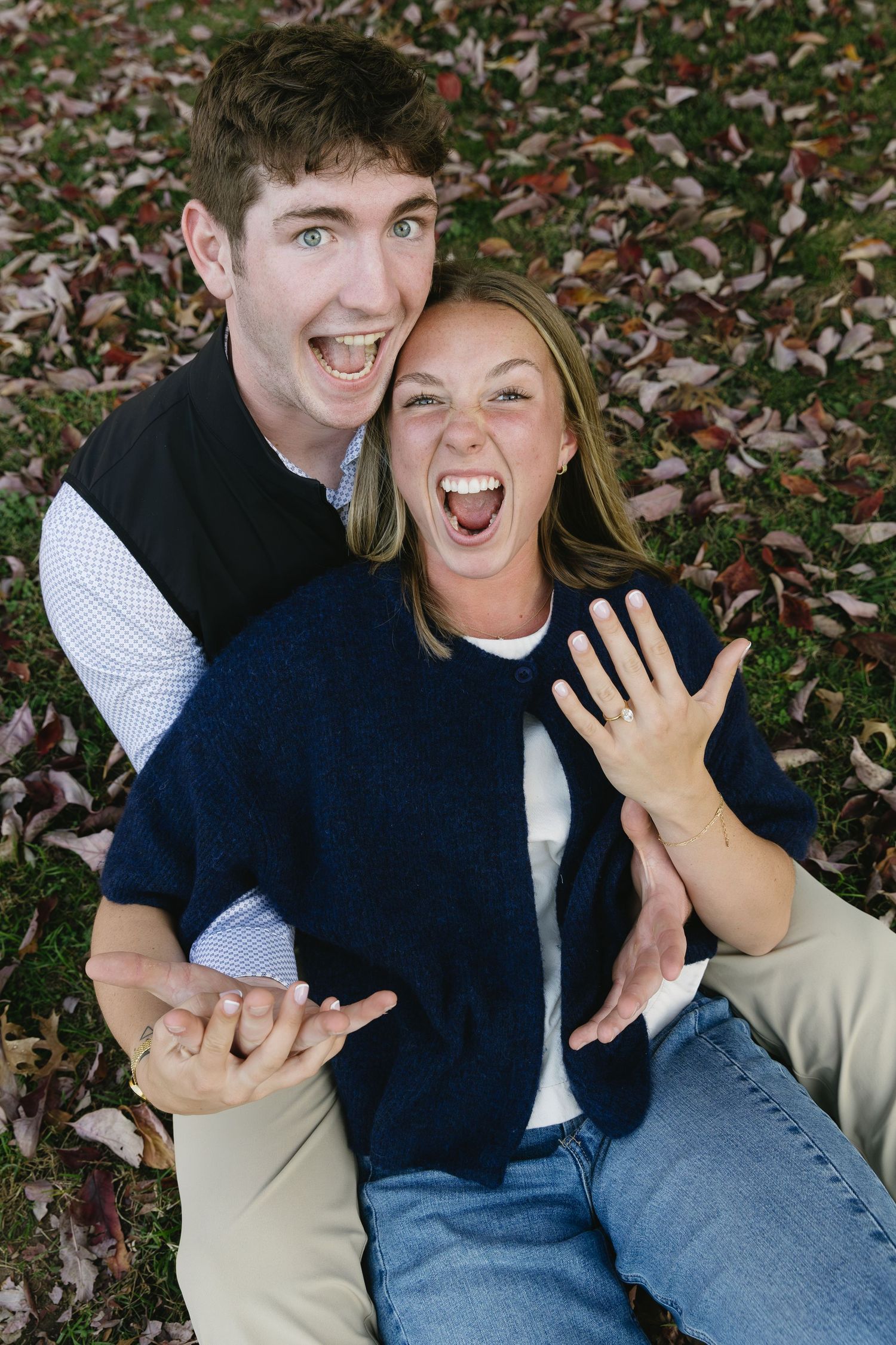 A couple sitting on fall leaves showing excitement and laughter while displaying a ring.