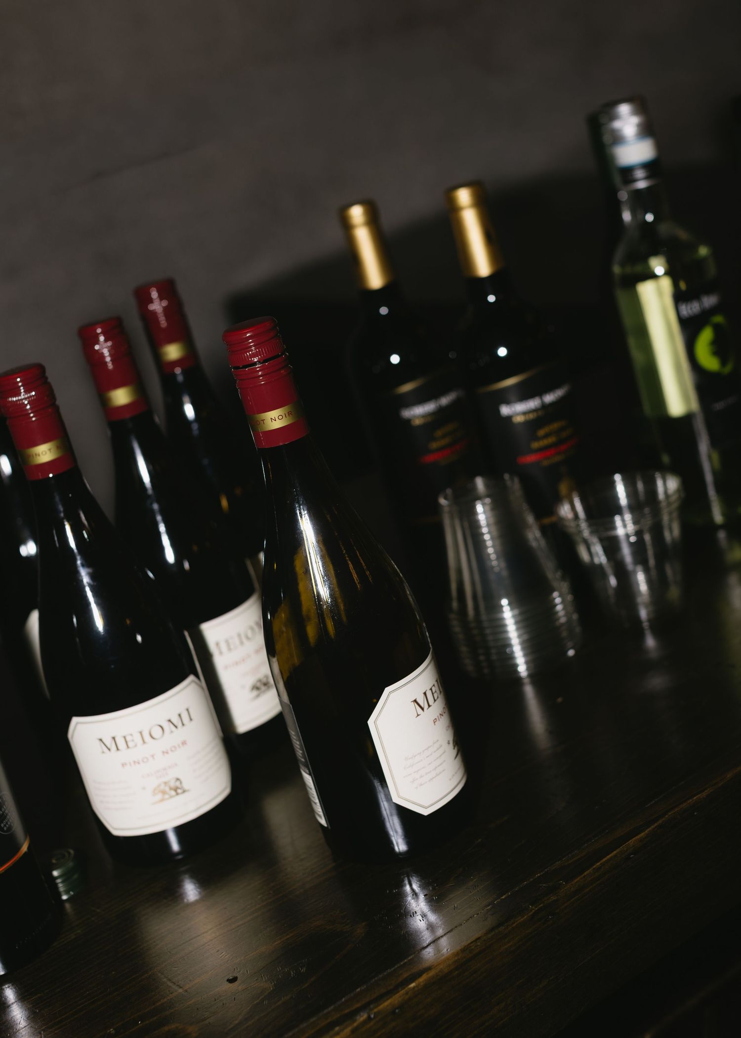 Collection of wine bottles lined up on a dark wooden surface in low lighting.