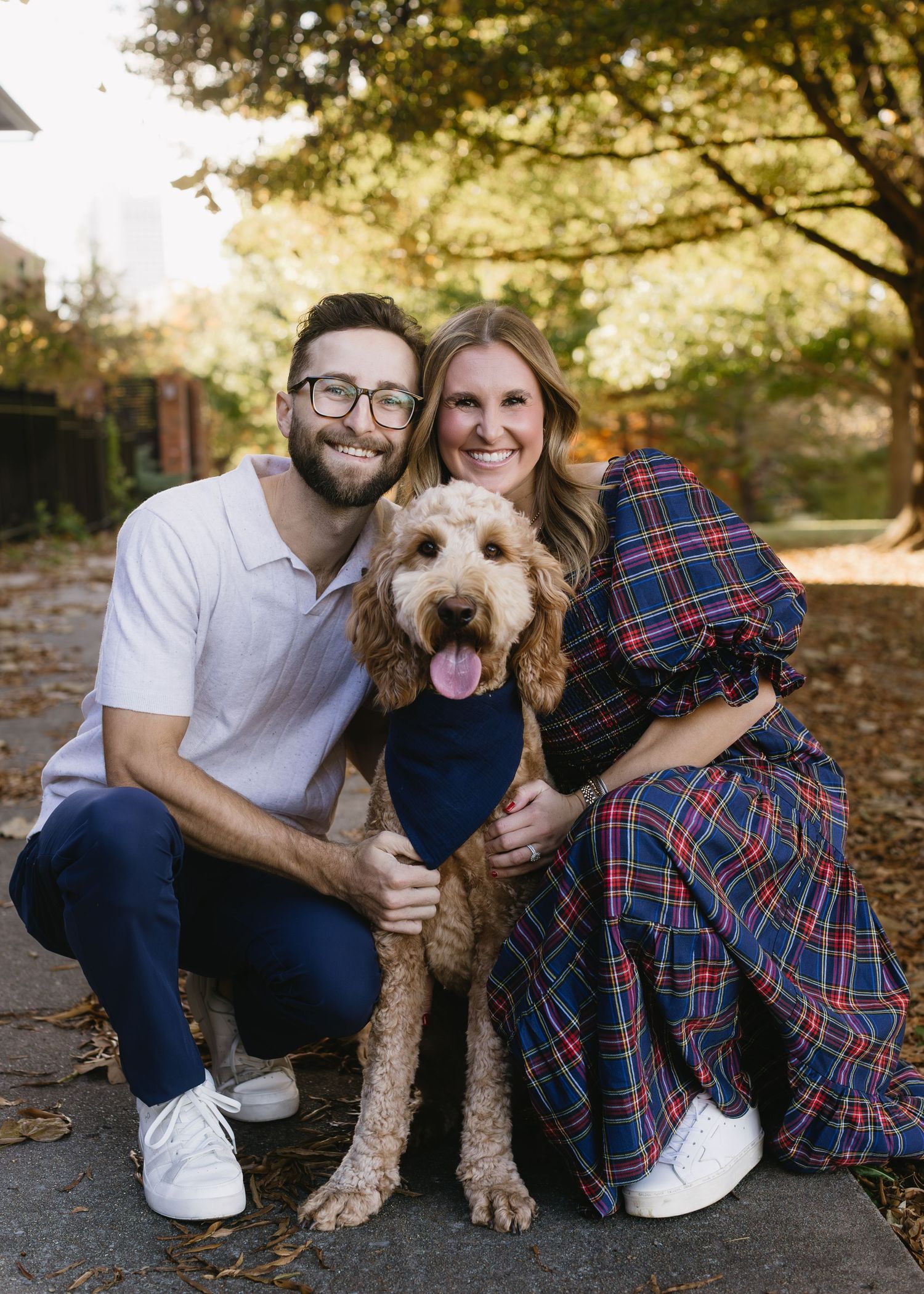 A couple and their goldendoodle dog pose together on fallen autumn leaves in an outdoor setting.