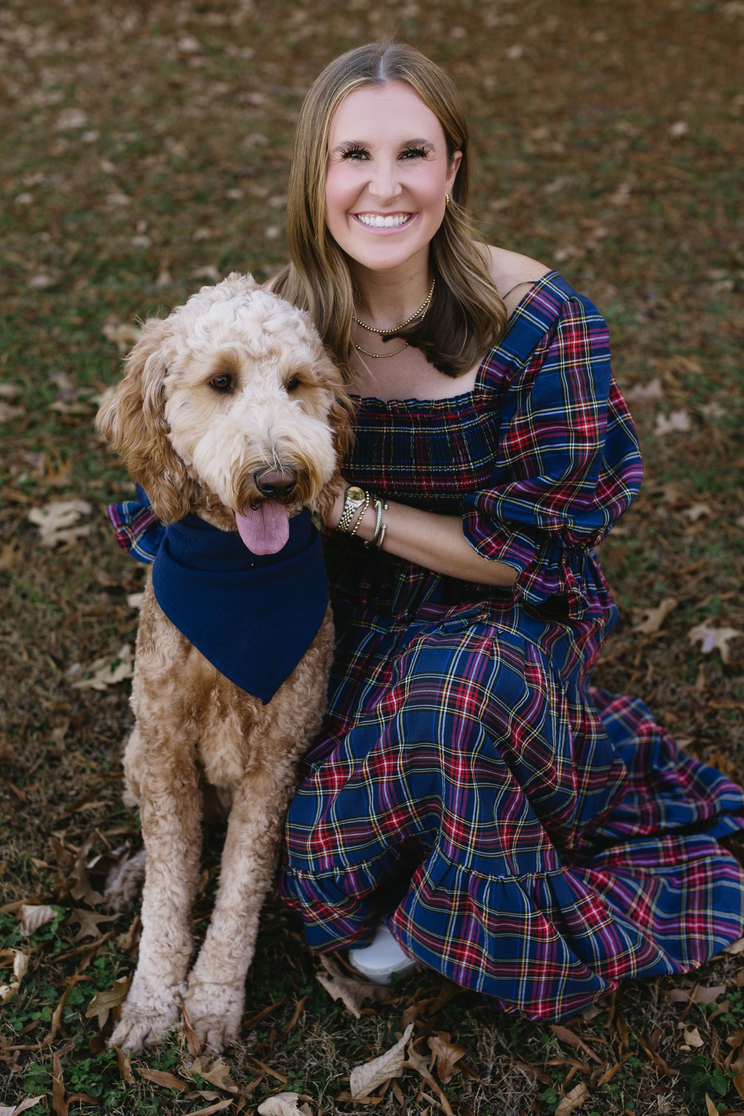 A goldendoodle dog wearing a navy bandana poses with someone in a plaid dress outdoors.