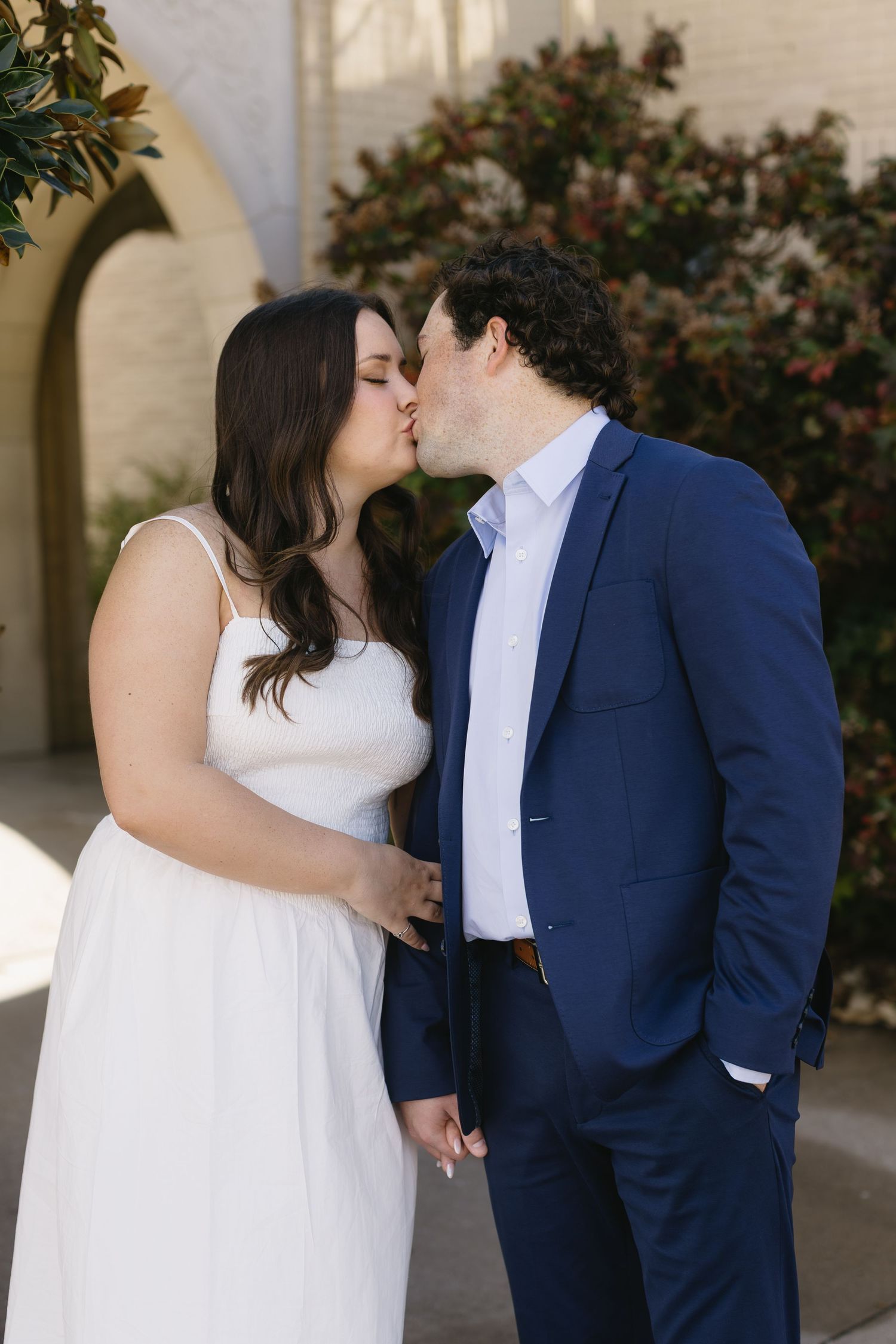 A romantic moment between a couple in formal wear standing against a white building exterior.