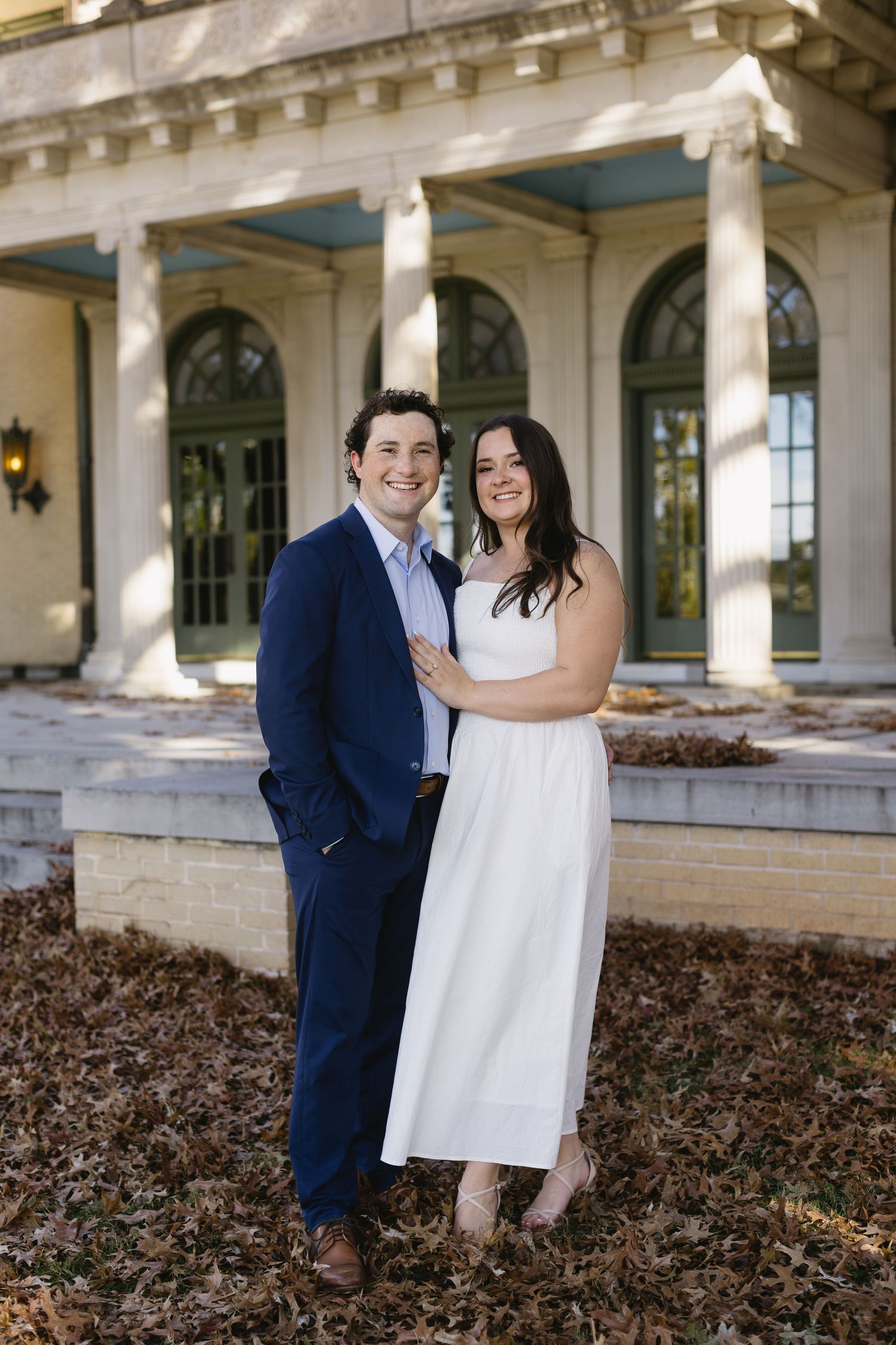 A couple poses in formal attire in front of a classical columned building entrance.