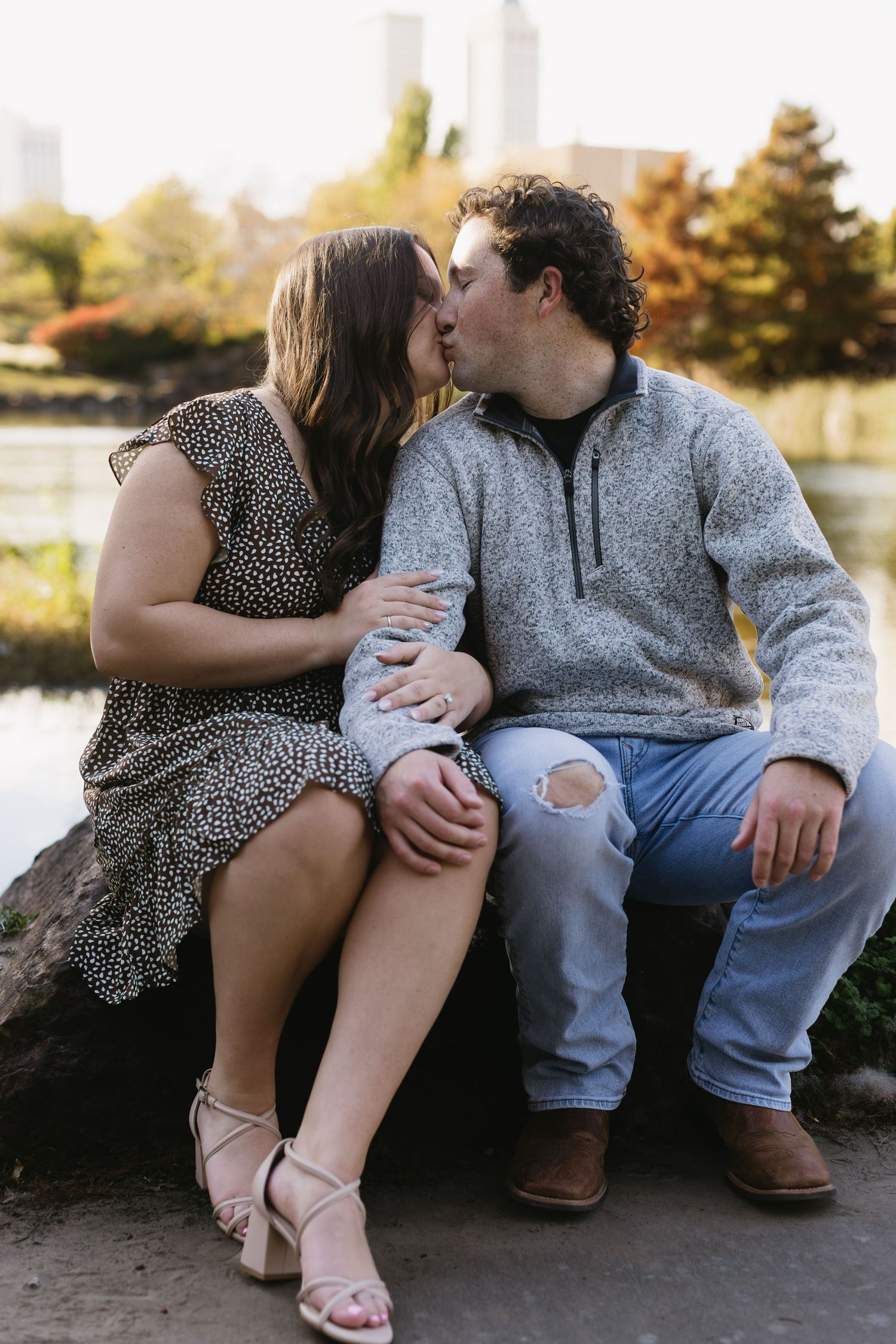 A couple in casual attire shares a romantic moment while sitting together on a stone ledge near a lake at sunset.