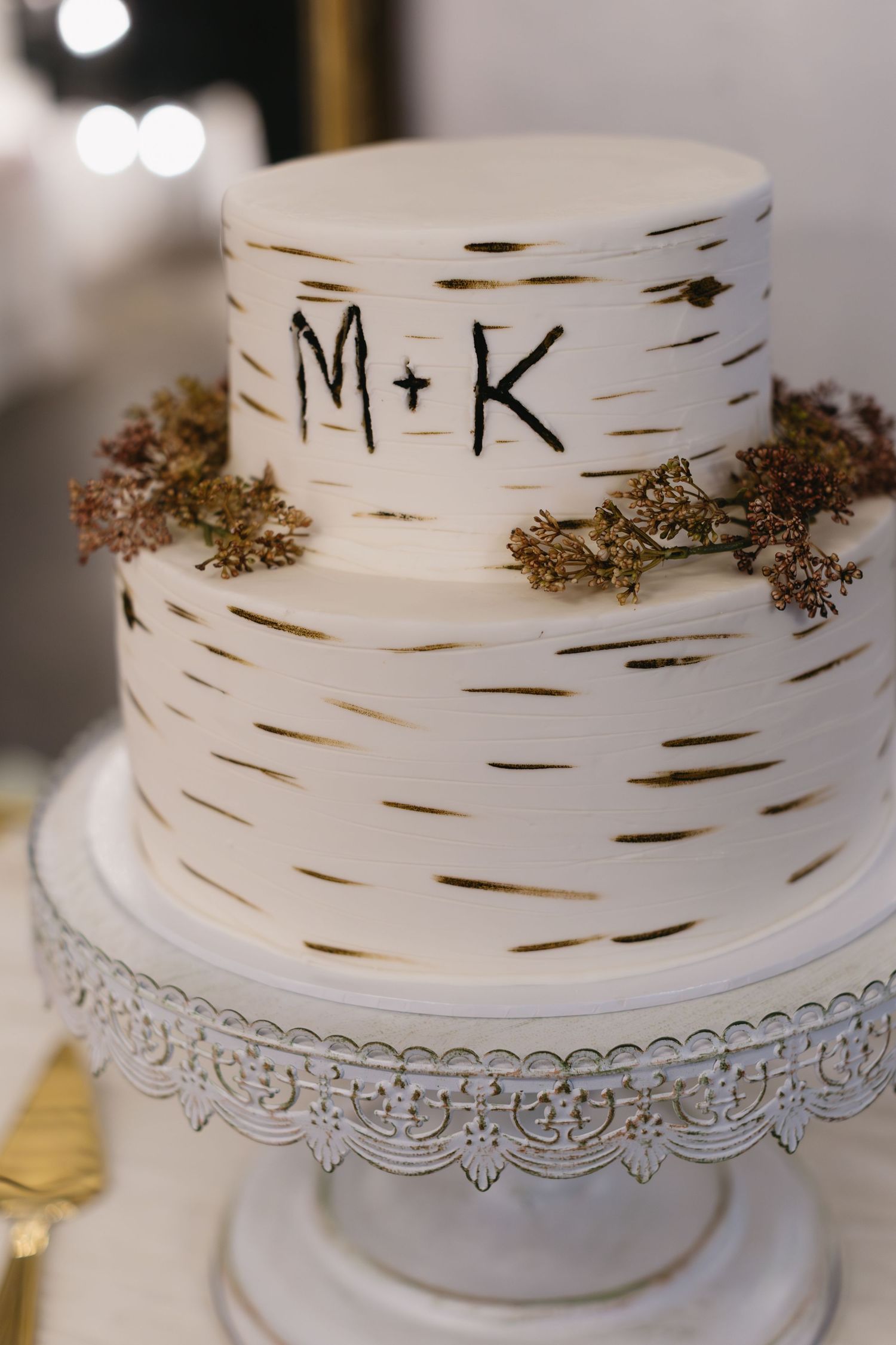 White two-tier wedding cake with birch bark design and initials M+K on an ornate silver cake stand.