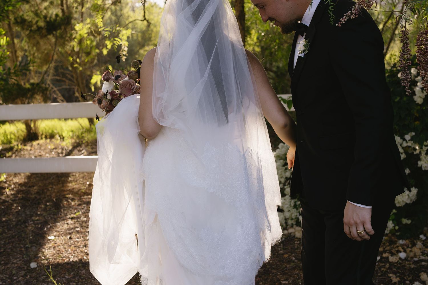 A bride in a white wedding dress and veil stands with her groom in a garden setting during their wedding ceremony.