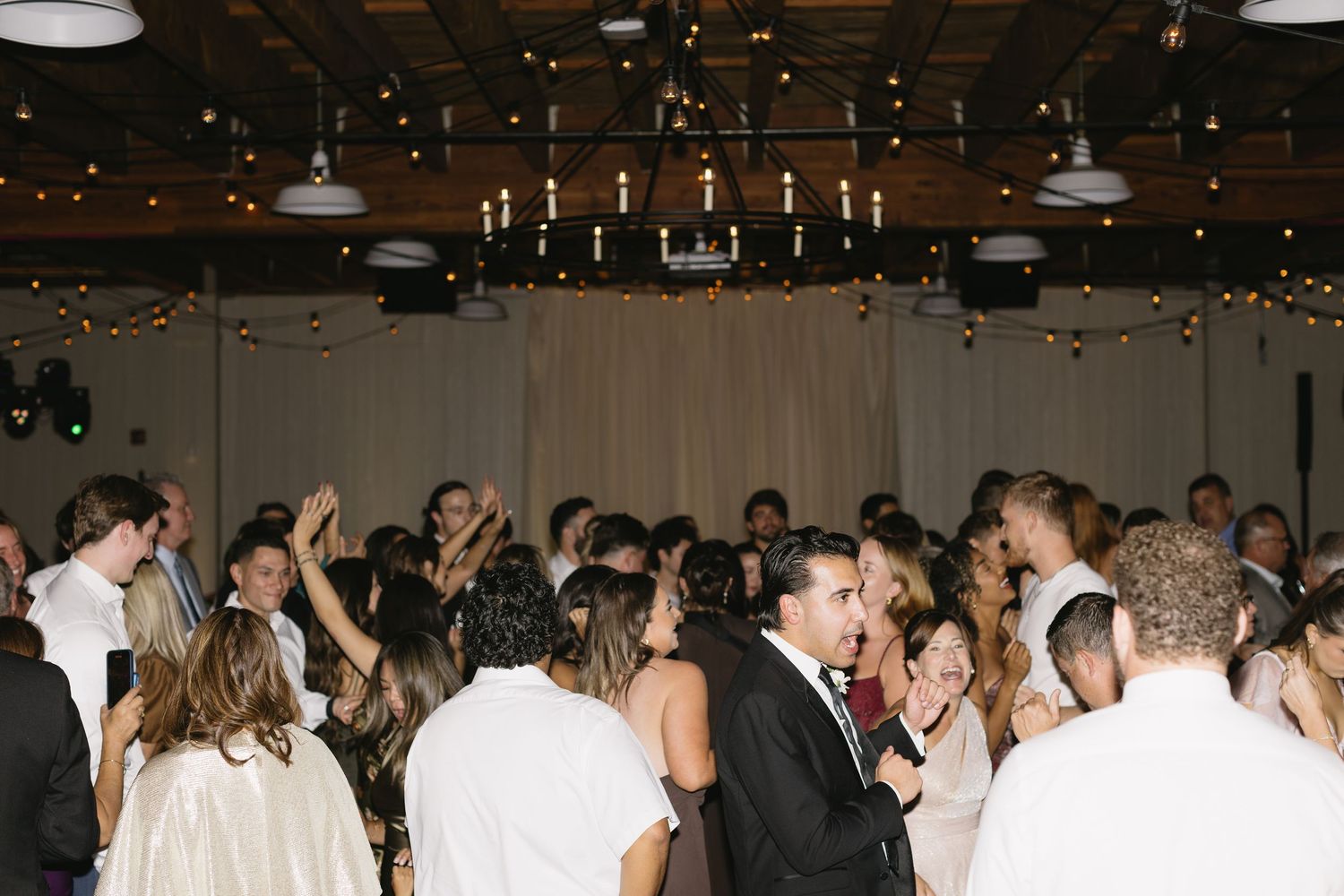 Wedding reception crowd dancing under string lights in industrial venue.