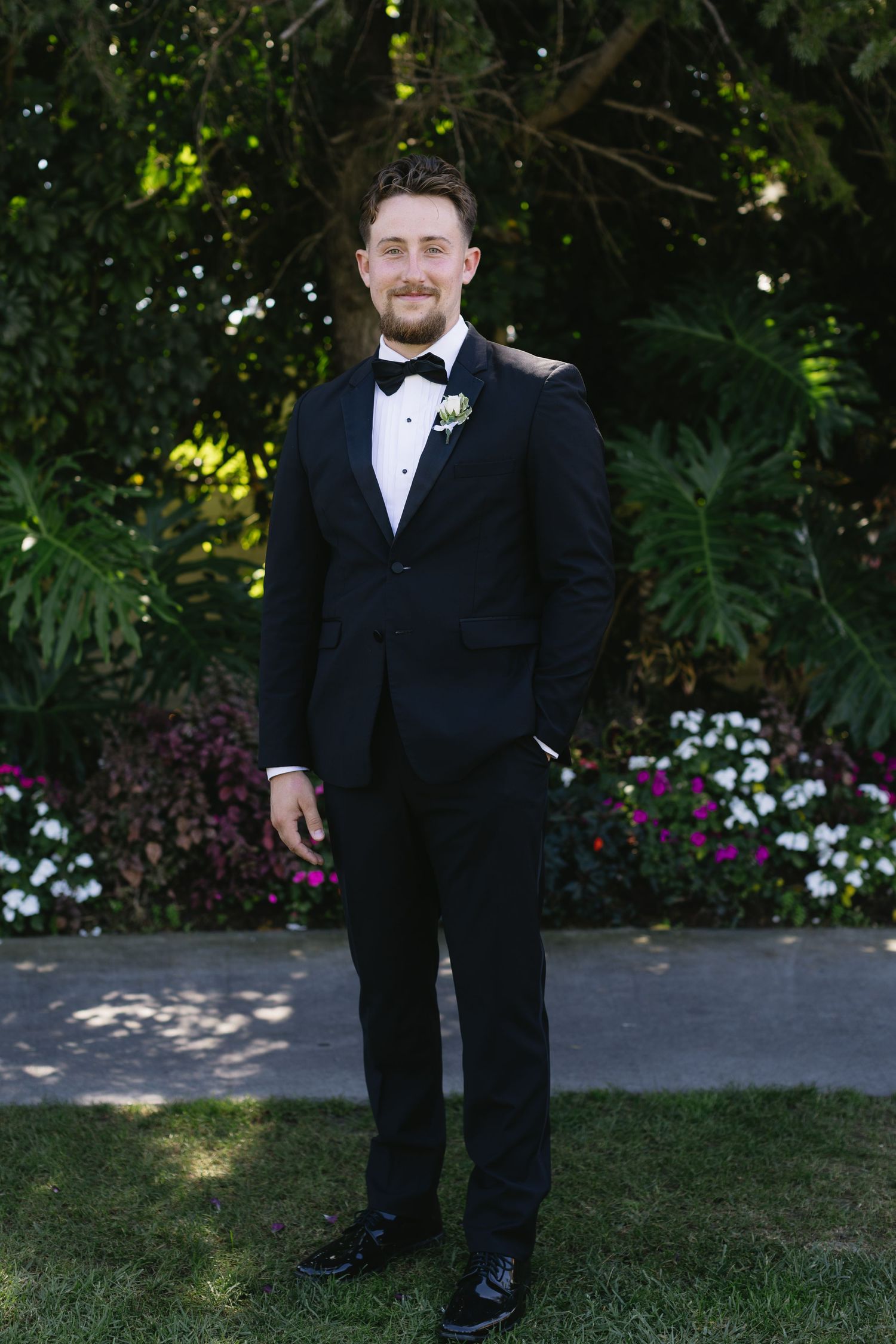 Groomsmen in black tuxedos and bow ties pose outdoors in a garden setting.