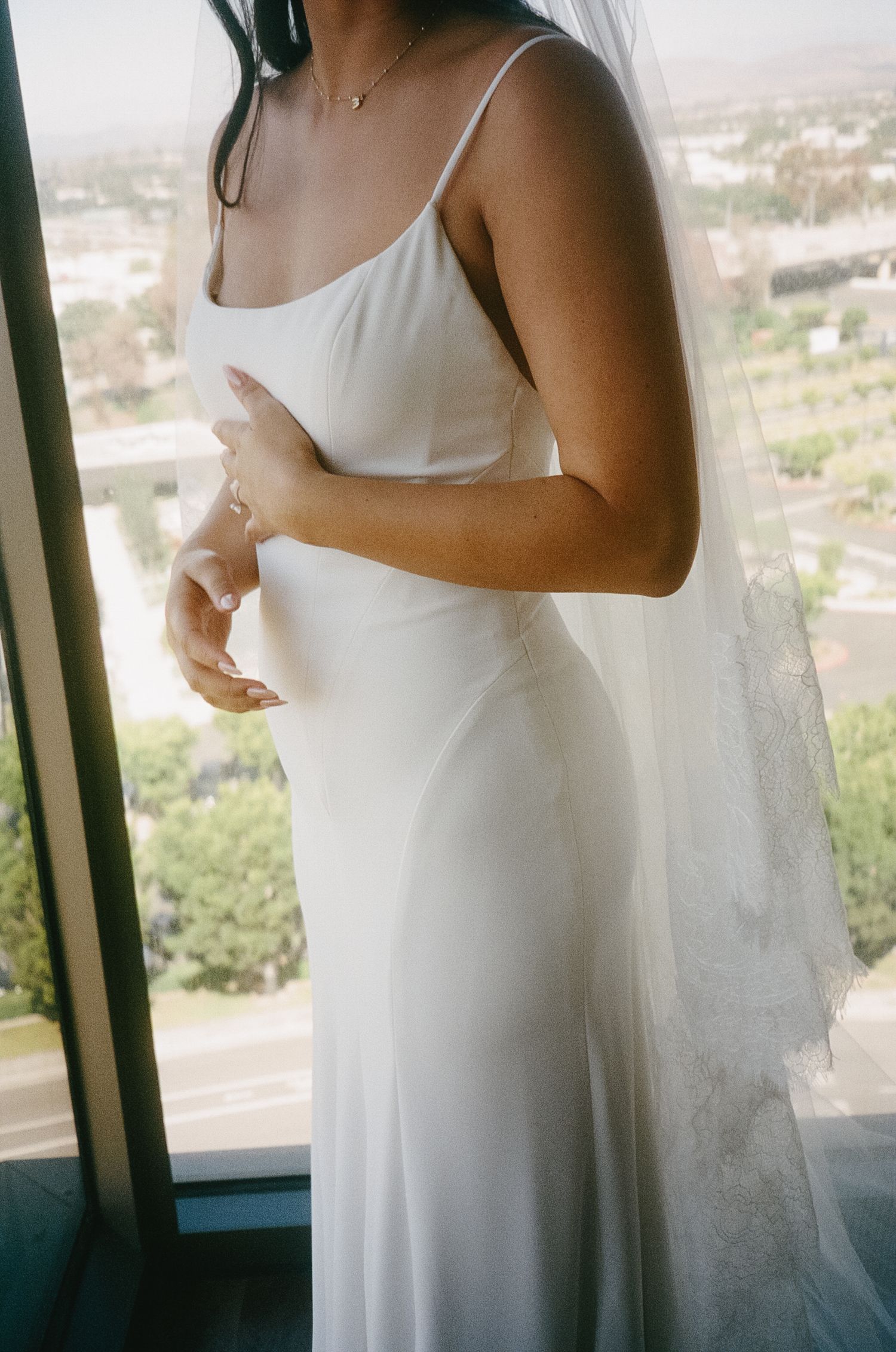 Close-up of bride in simple white wedding dress standing by window with city view.