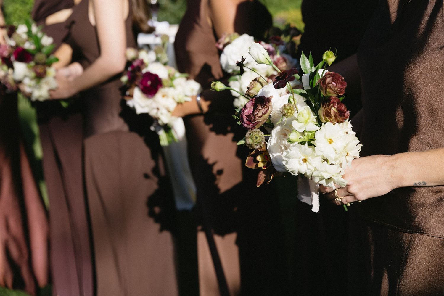 Row of bridesmaids in brown dresses holding white and pink floral bouquets.