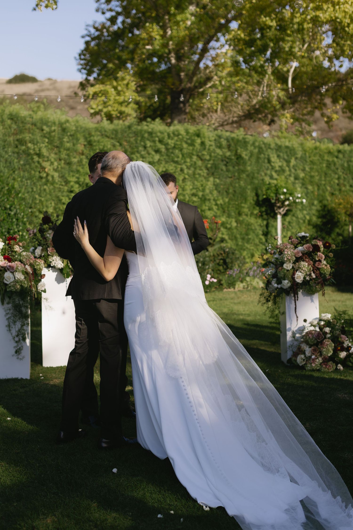 Flowing white wedding dress and veil trail behind couple during outdoor garden ceremony.