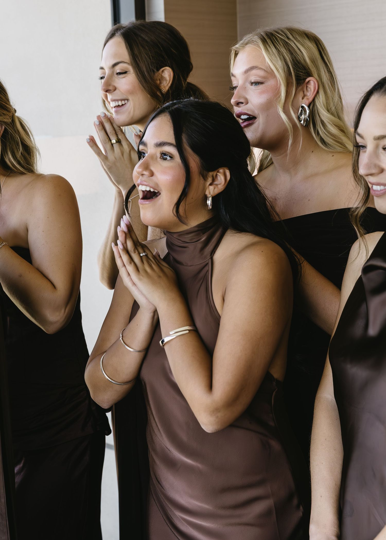 Bridesmaids in brown dresses share excited reactions during wedding preparations.