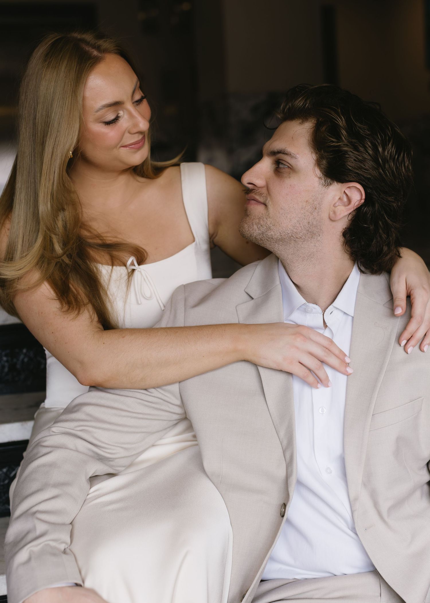 A romantic couple in light-colored formal wear share an intimate moment while sitting together indoors.