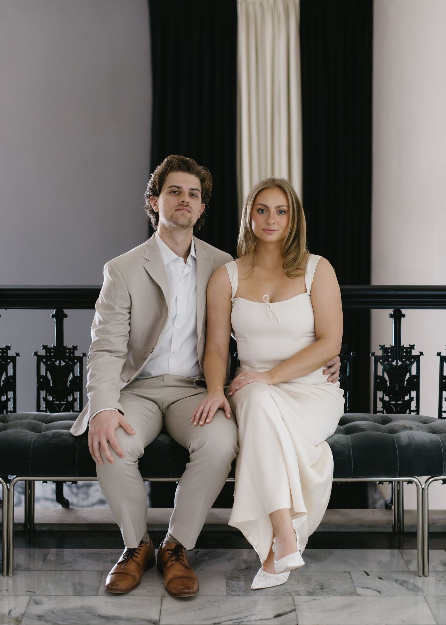A couple sits together on a black bench wearing matching formal attire in light colors.