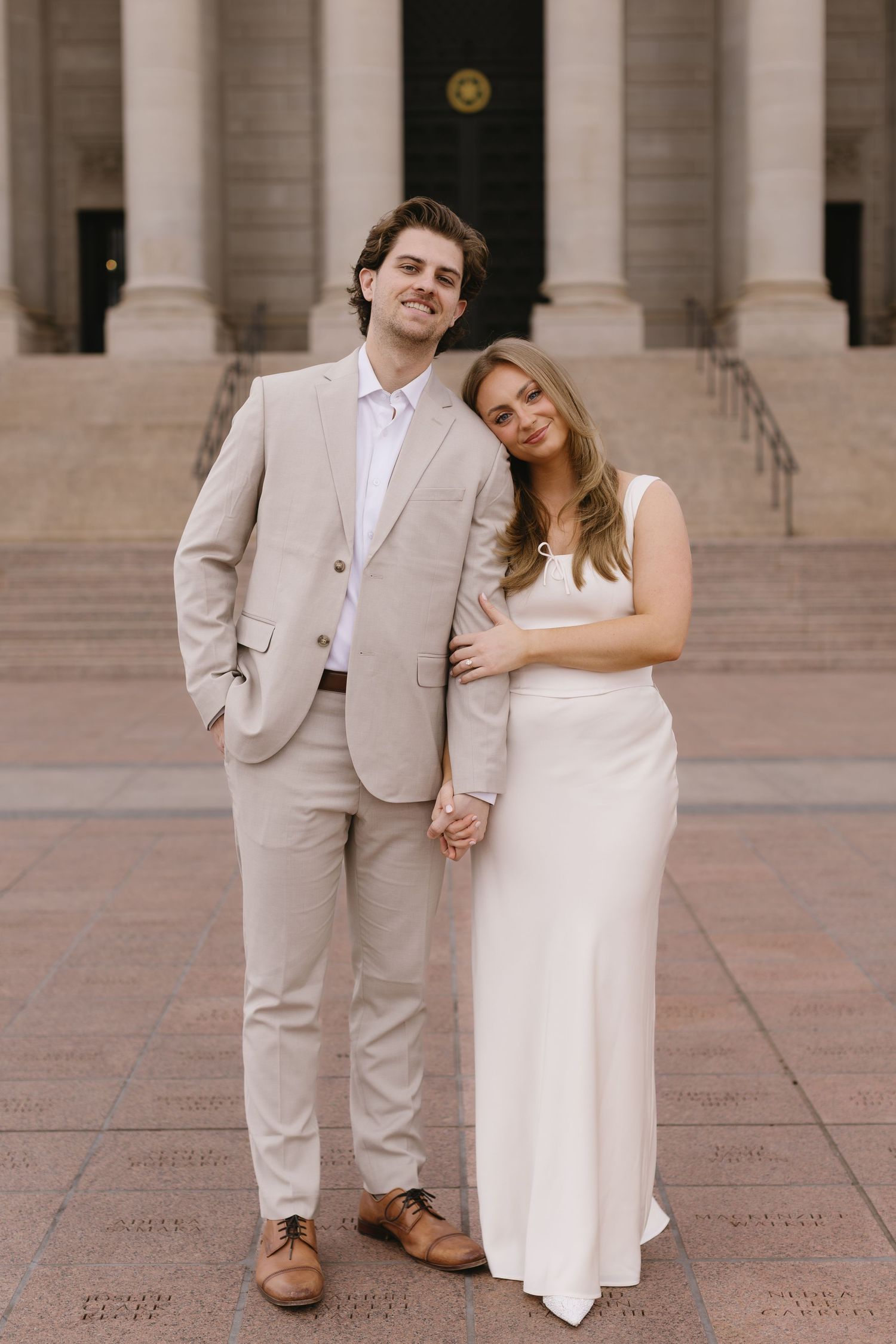 A romantic couple in coordinating beige and white outfits poses together on stone steps of a grand building.