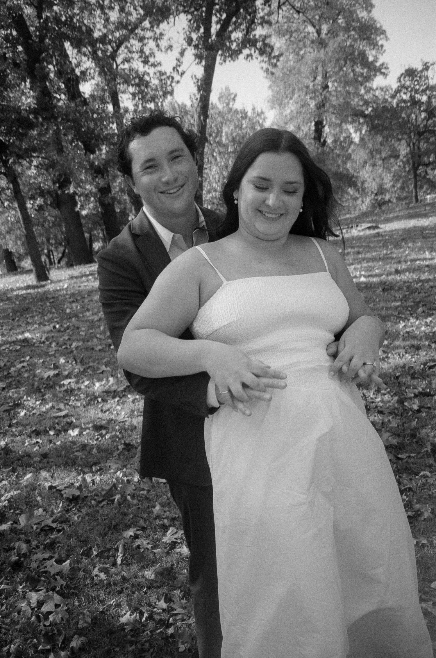 A black and white wedding photo of a couple embracing outdoors in a wooded setting.