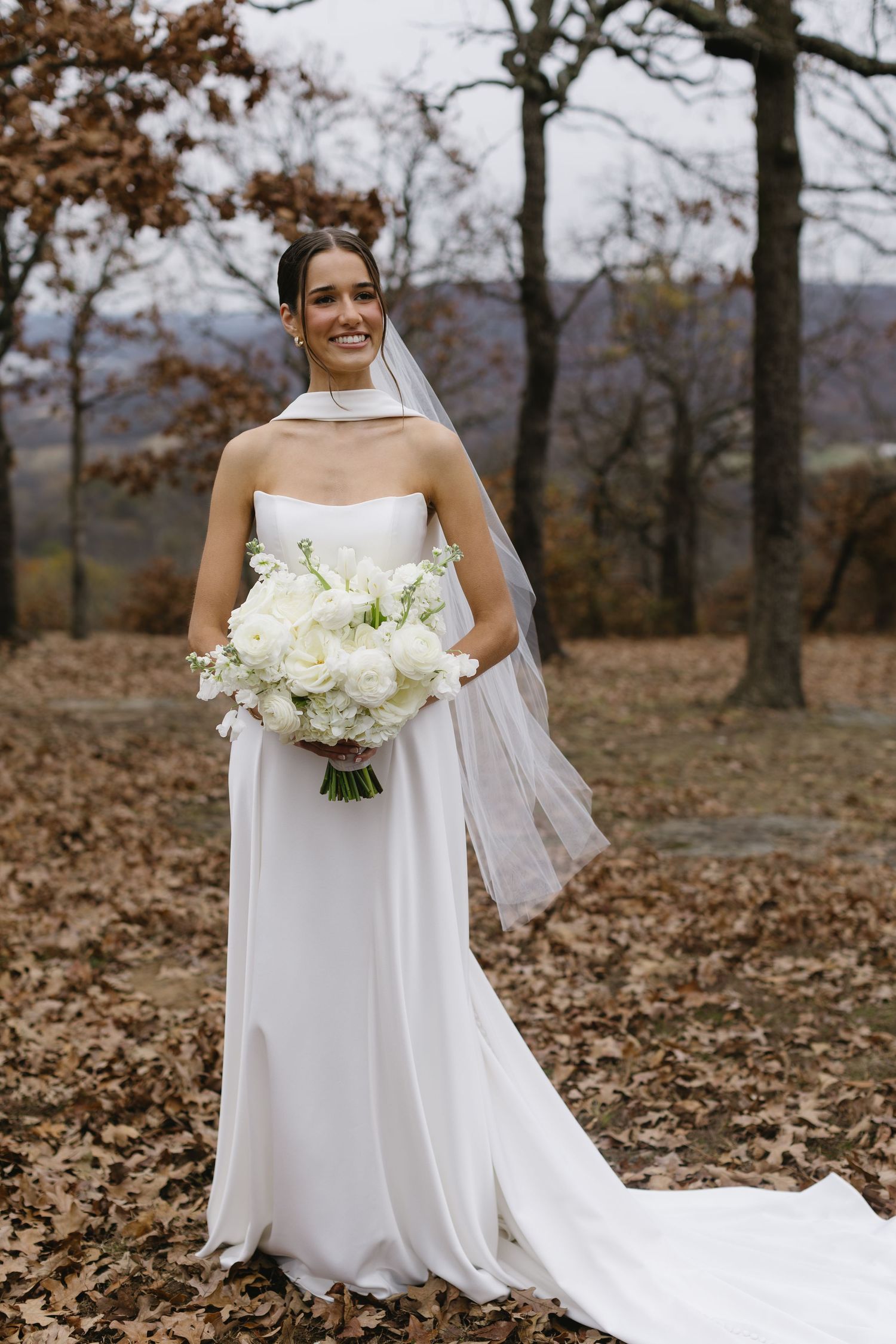 A bride poses outdoors in autumn holding a white flower bouquet against a mountain backdrop.