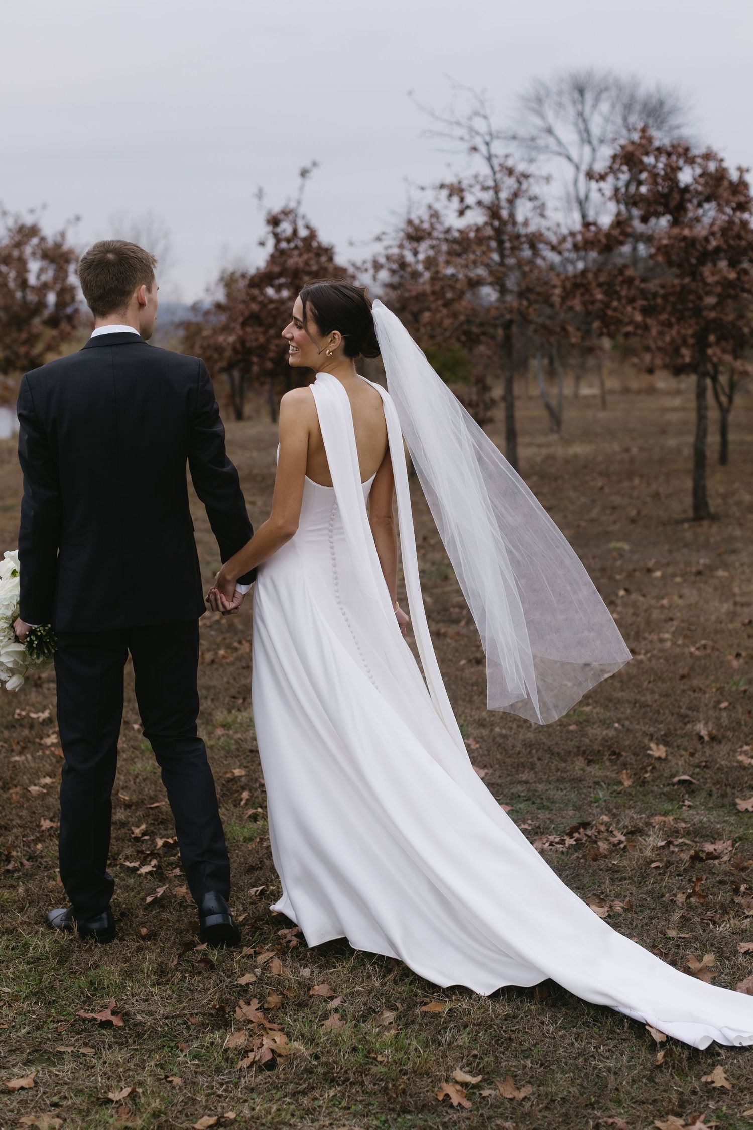 Wedding couple walking through autumn landscape with flowing white dress train.
