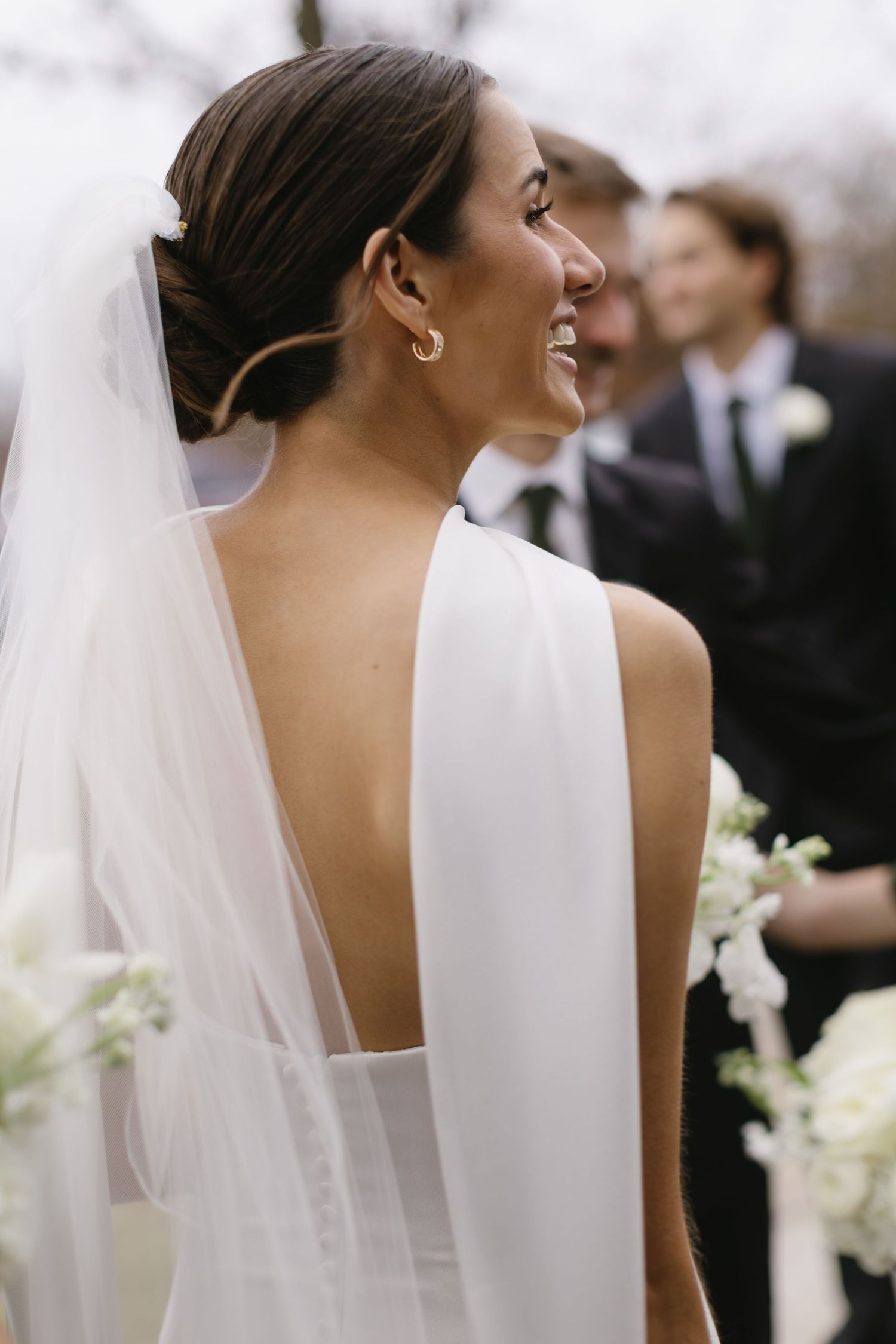 A bride in a sleek white dress and veil smiles during her wedding ceremony.