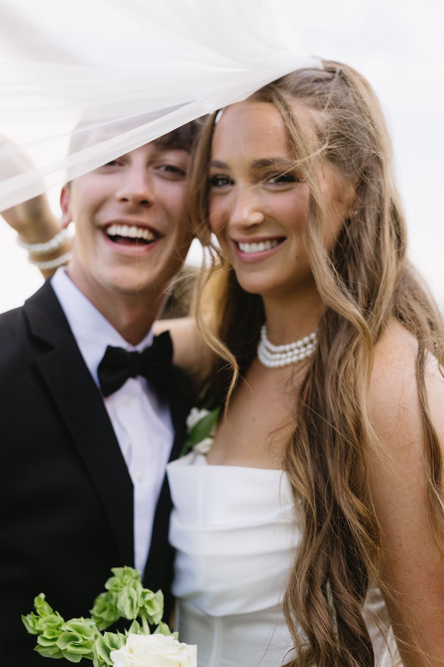 A joyful wedding couple sharing a candid moment with their faces close together, the bride wearing a white gown and pearl necklace.