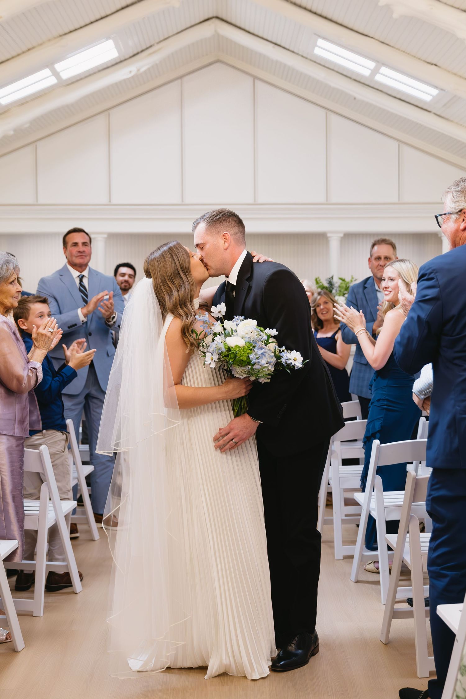 A wedding ceremony kiss in a white chapel with guests applauding.