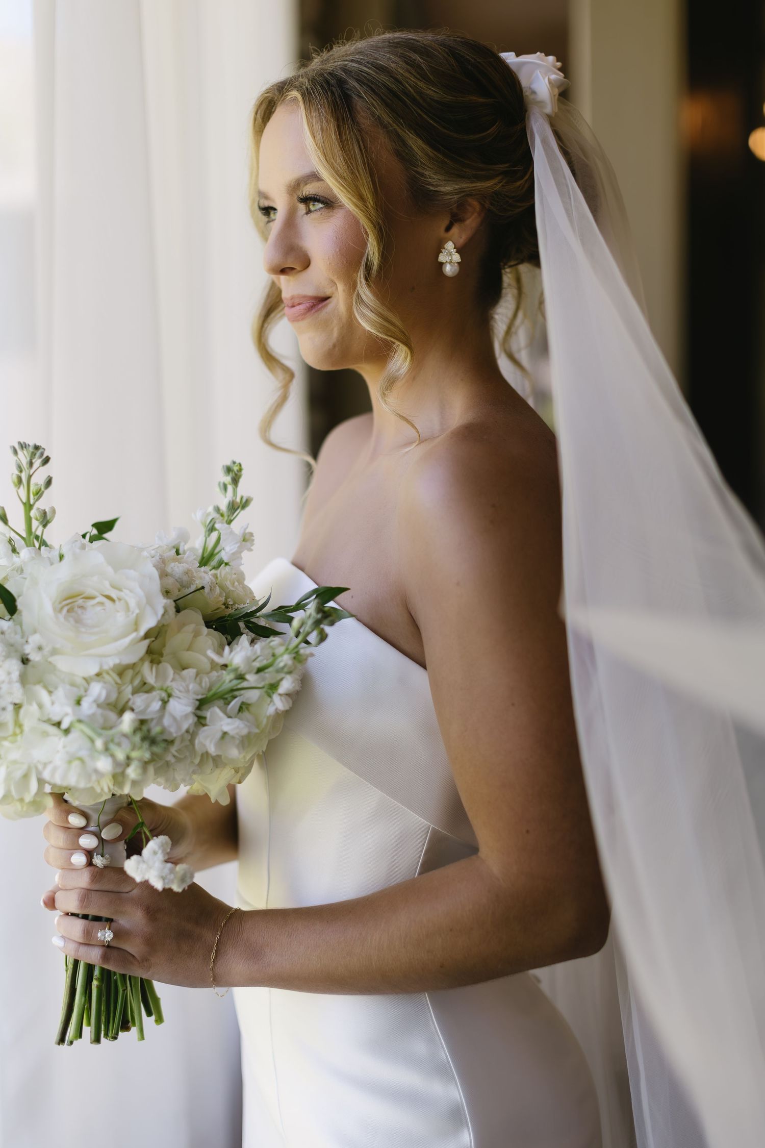 Bride in white wedding dress holds elegant white floral bouquet while standing near window in natural light.