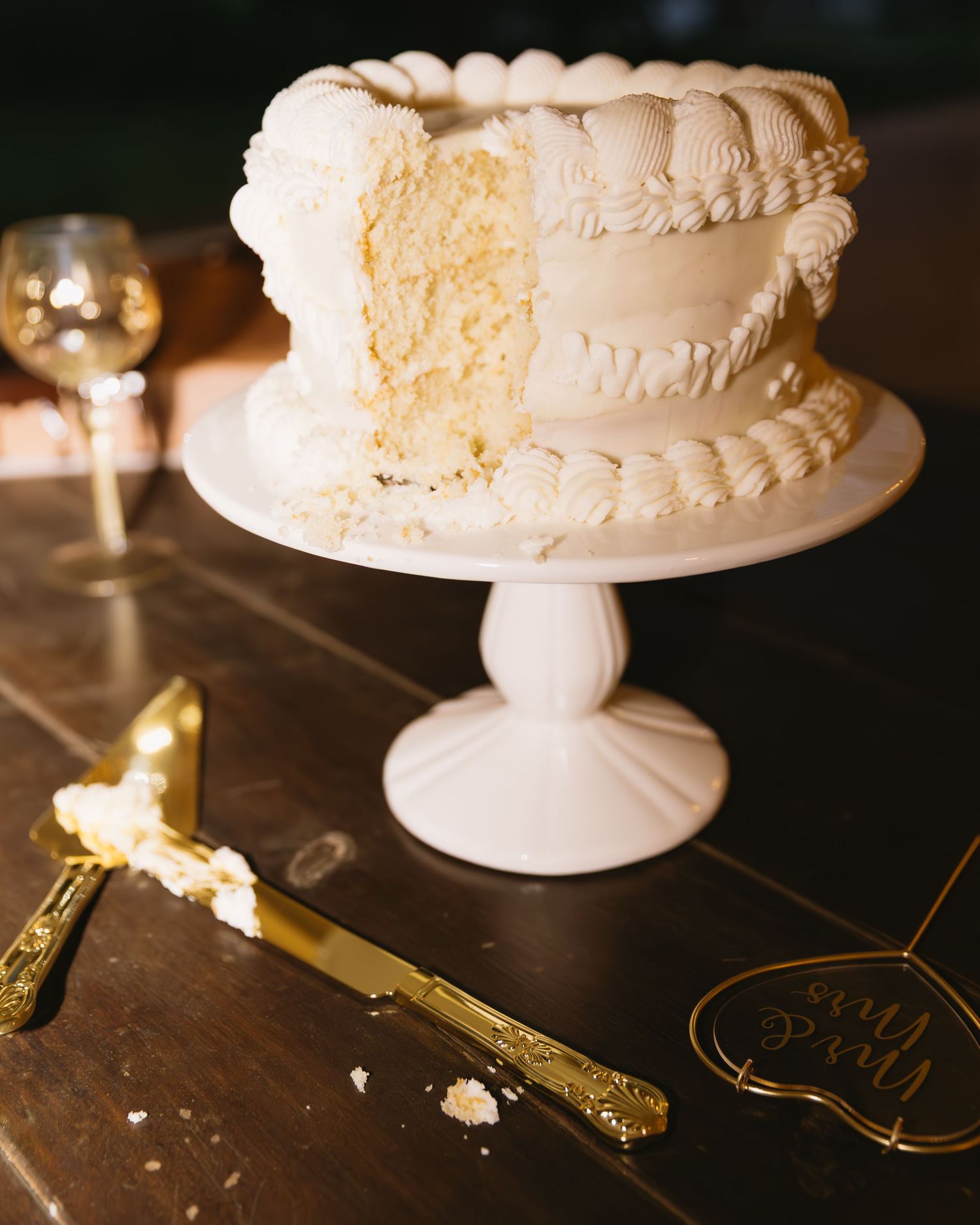 Elegant white wedding cake on a pedestal stand with gold serving utensils on a dark wooden table.