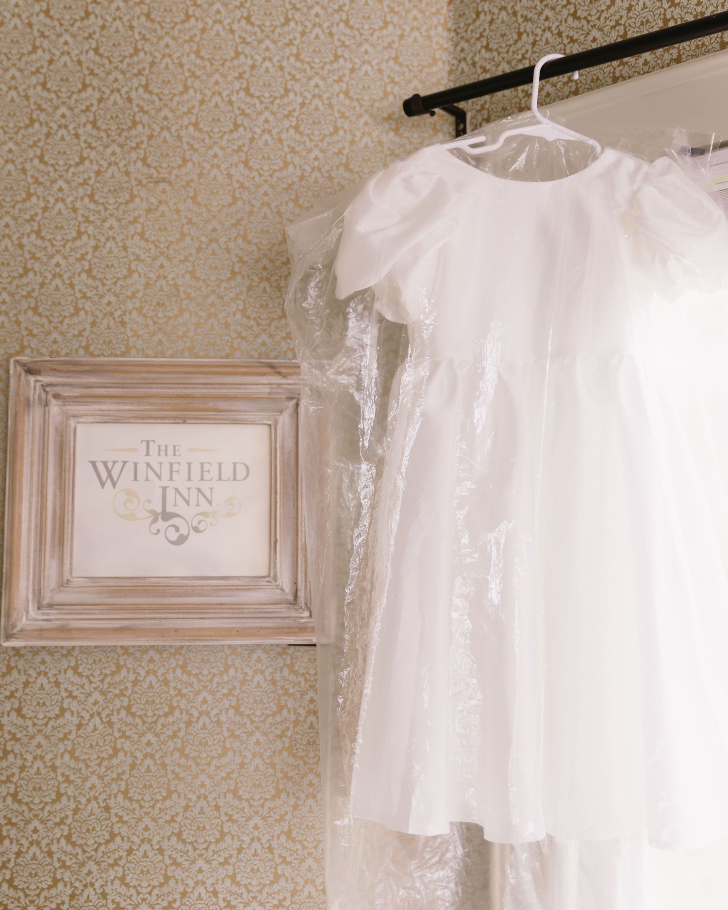A delicate white christening gown hanging beside a Winfield Inn sign on a textured wallpaper background.