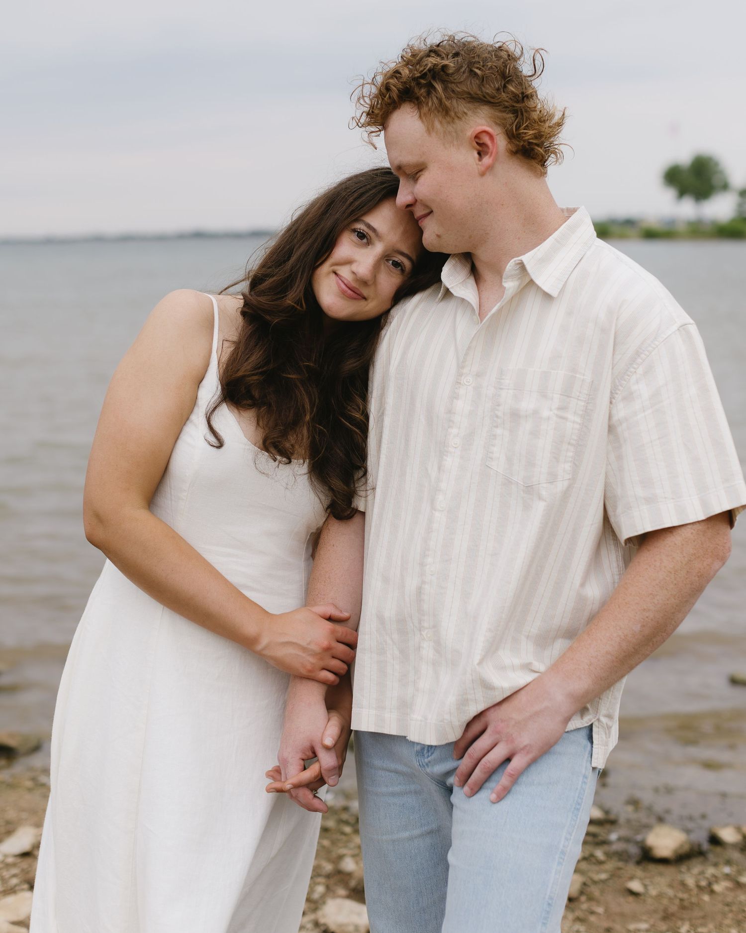A romantic couple poses together by the waterside wearing coordinating white and cream outfits.
