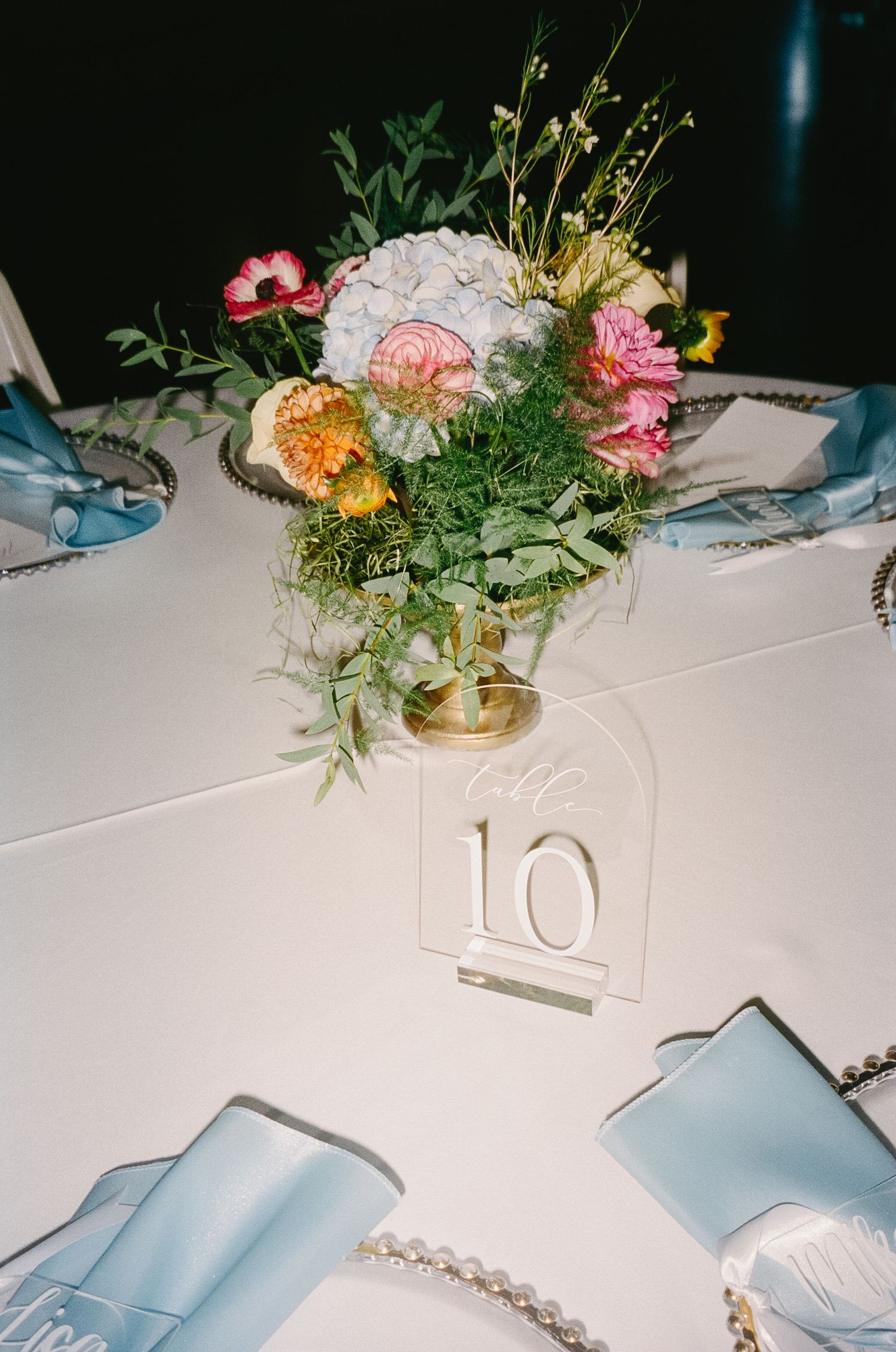 An elegant floral centerpiece with pink and white flowers in a gold vase sits on a white tablecloth with table number 10.