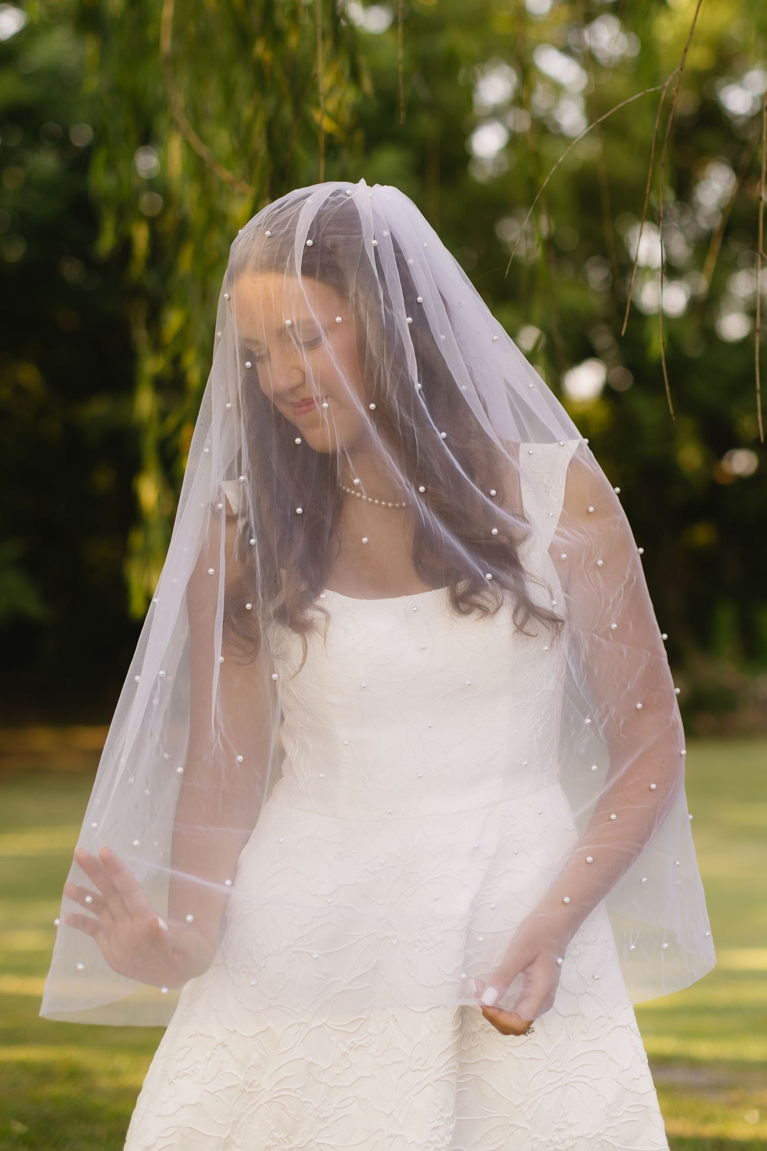 A bride wearing a pearl-adorned veil poses under natural outdoor lighting.