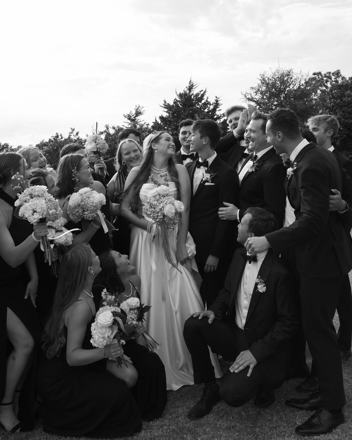 Wedding party gathered for a candid black and white group photo with bouquets and formal attire.
