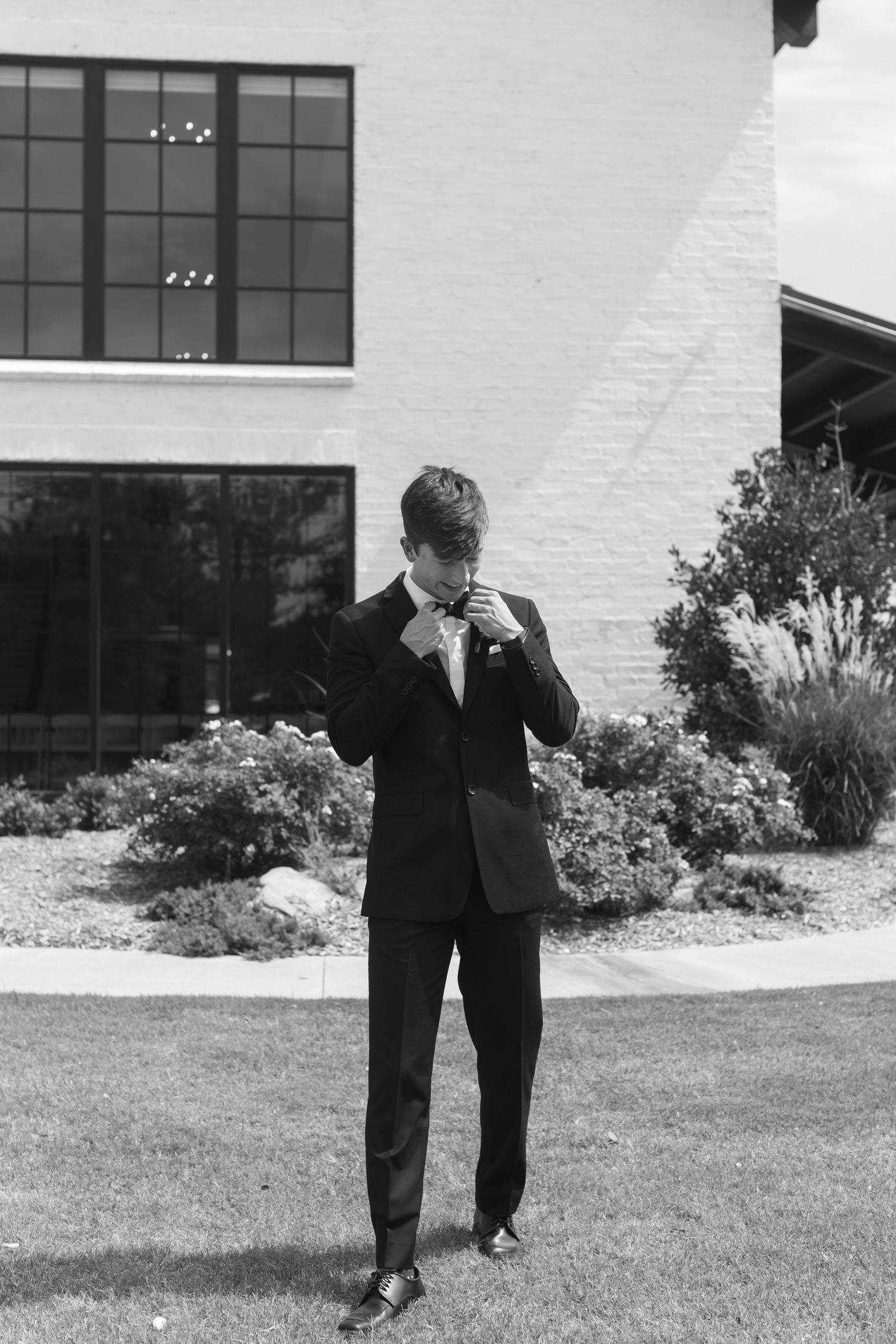 Formal suited figure adjusting tie while standing on landscaped lawn in black and white.