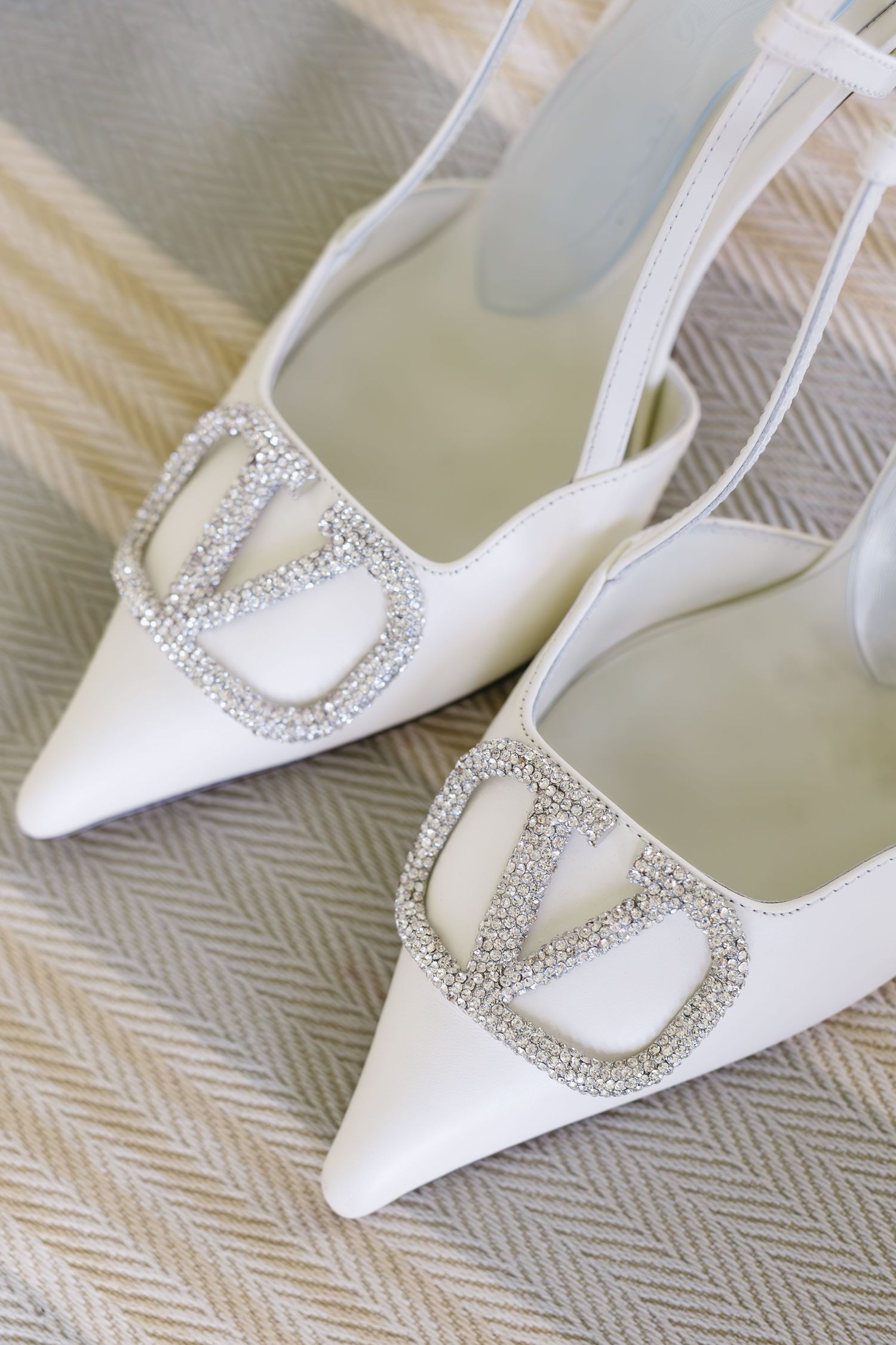 Elegant white bridal shoes with crystal embellishments on textured gray fabric.
