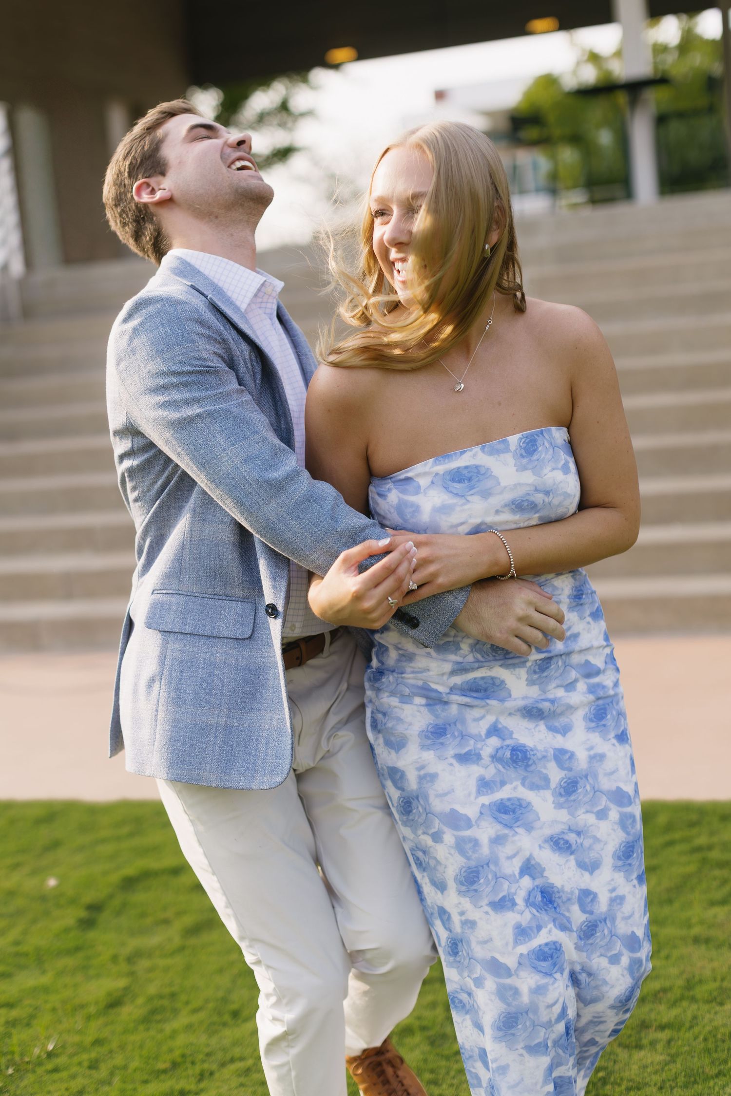 A couple embraces and laughs together outdoors, with one wearing a light blue dress and the other in grey.