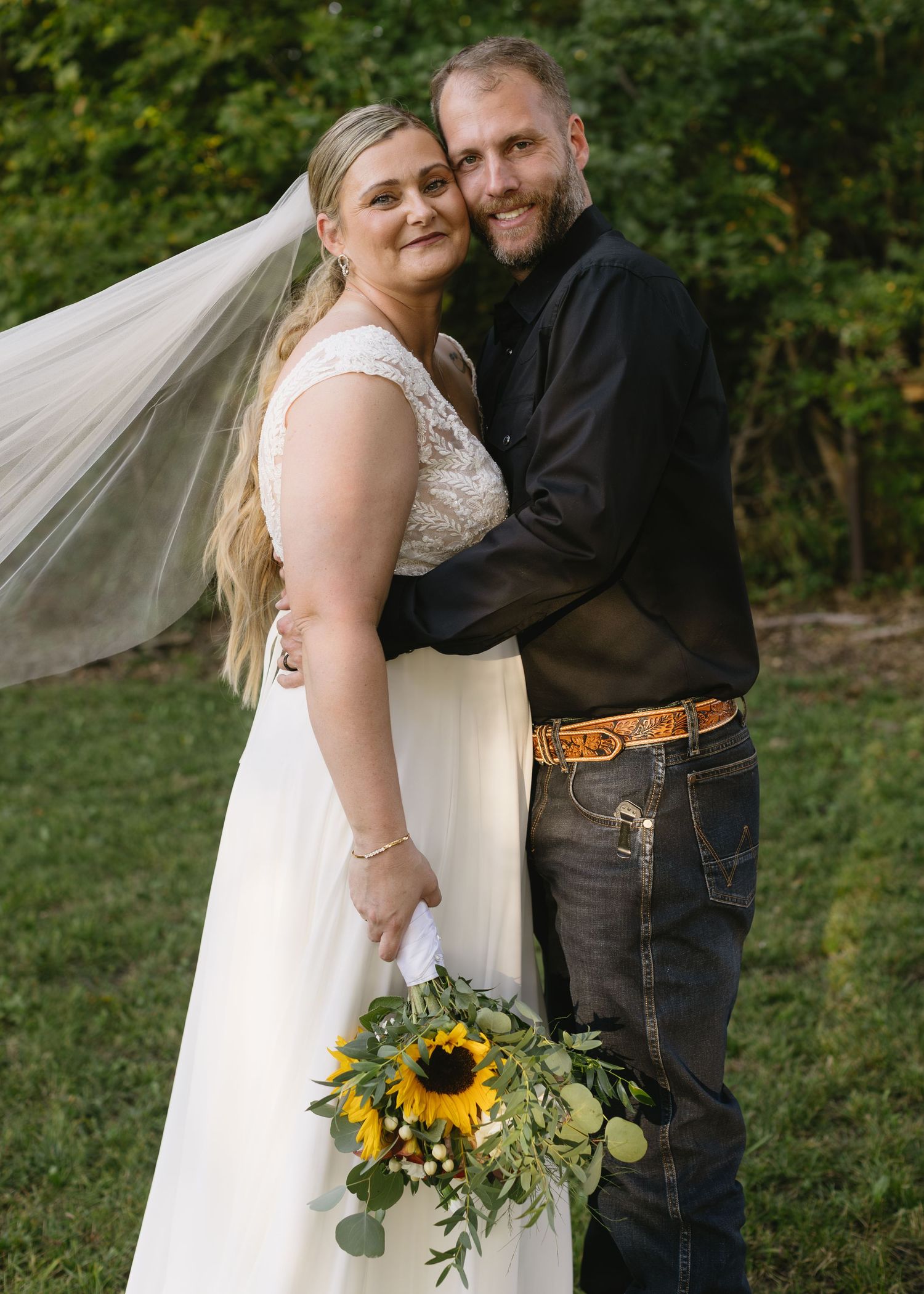 A couple poses outdoors at their wedding with sunflower bouquet and flowing white dress.