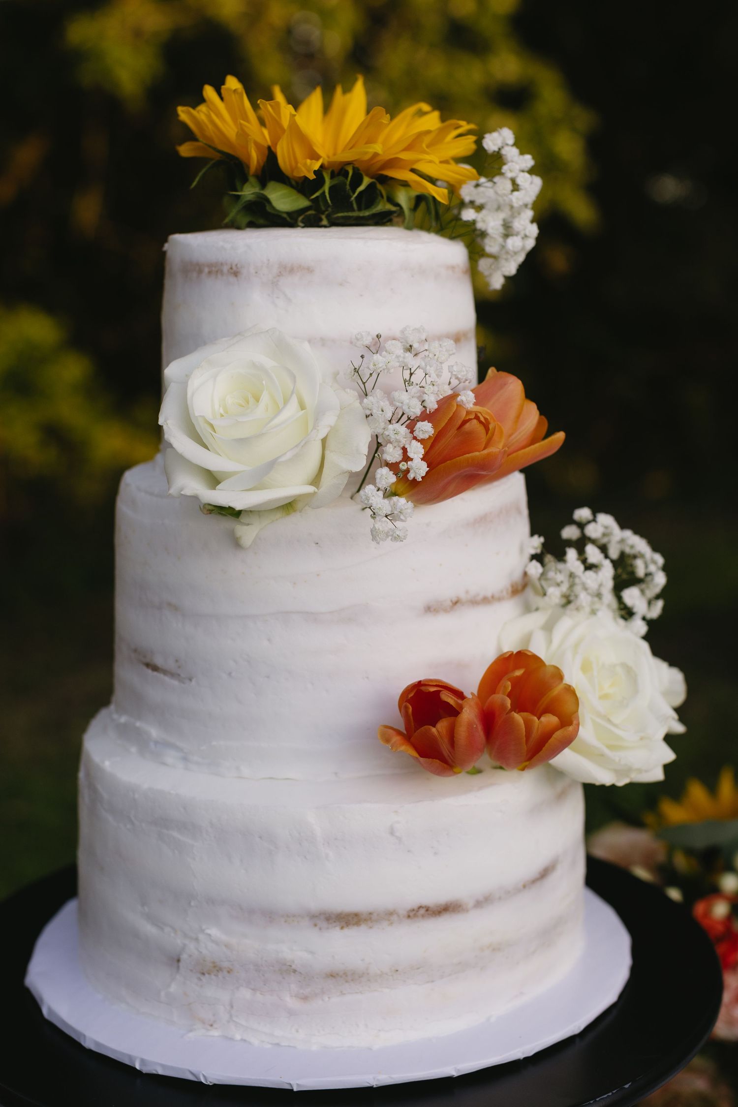 A rustic white wedding cake decorated with sunflowers, roses and baby's breath on a black cake stand.