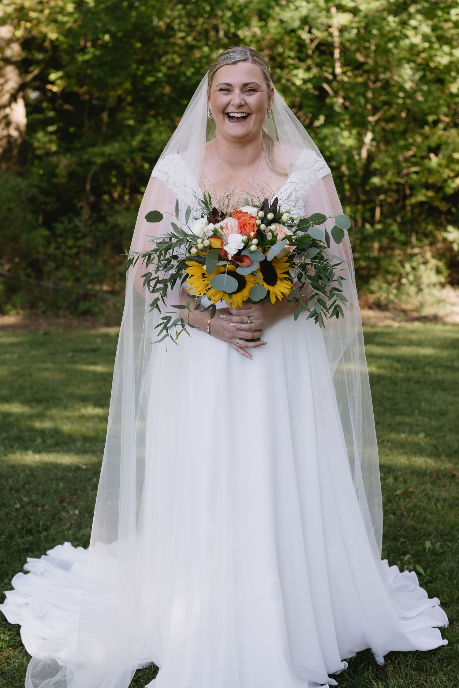A bride in a flowing white gown holds a vibrant sunflower bouquet in an outdoor garden setting.