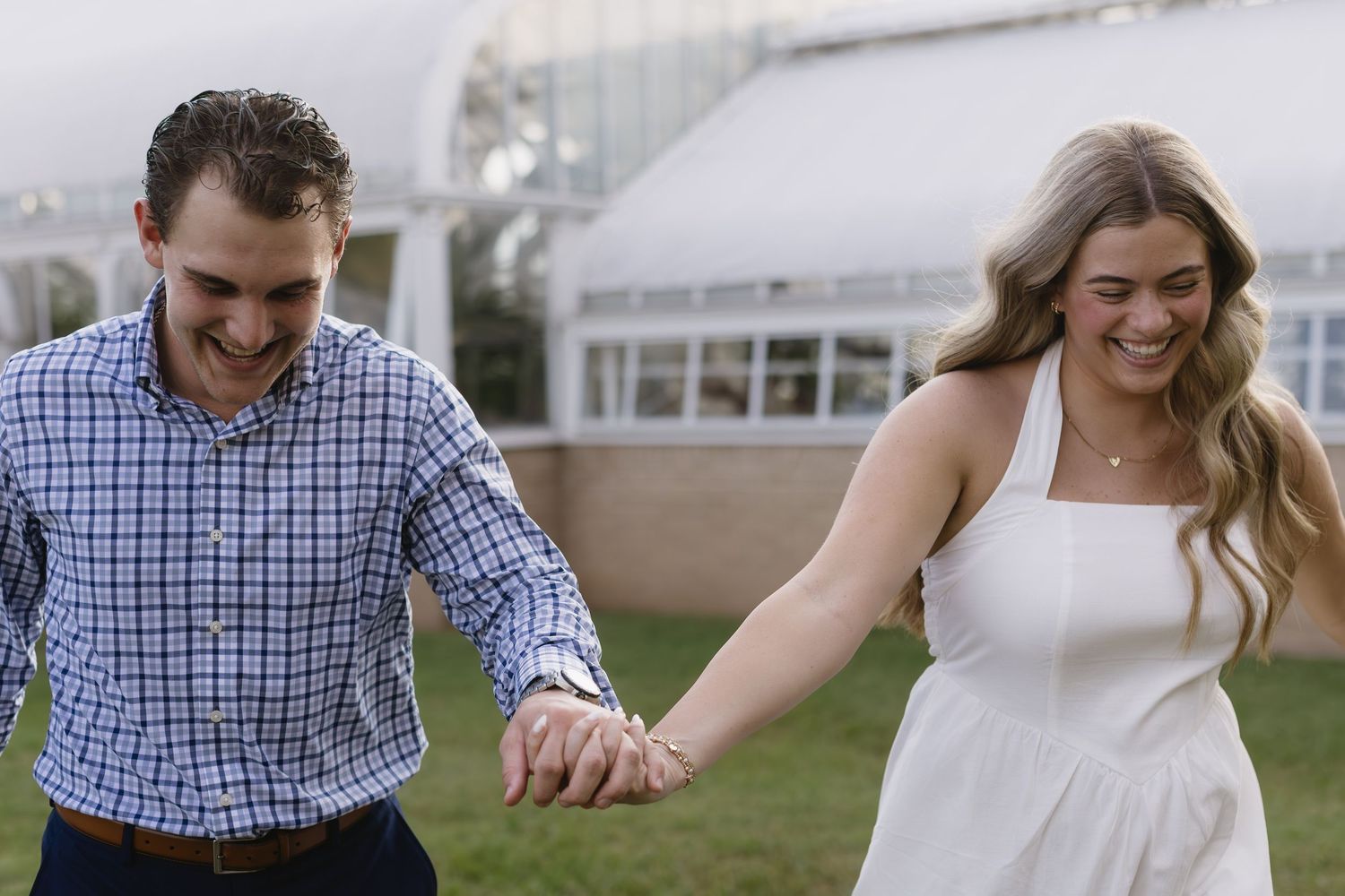 A couple holds hands and runs together outside, with one wearing a blue plaid shirt and the other in a white dress.