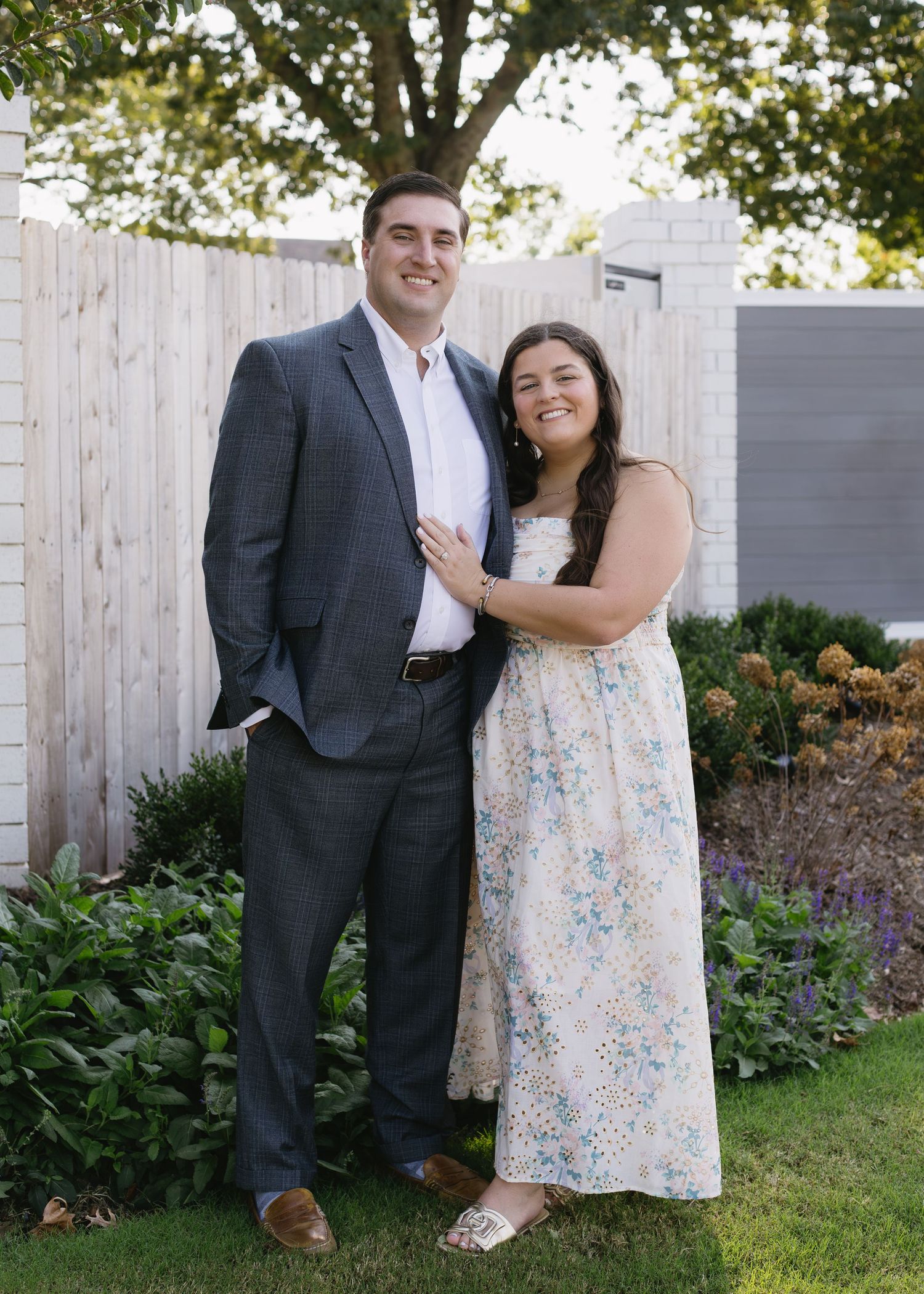 A romantic couple poses for photos in a garden with a wooden fence backdrop, wearing formal attire and a floral dress.