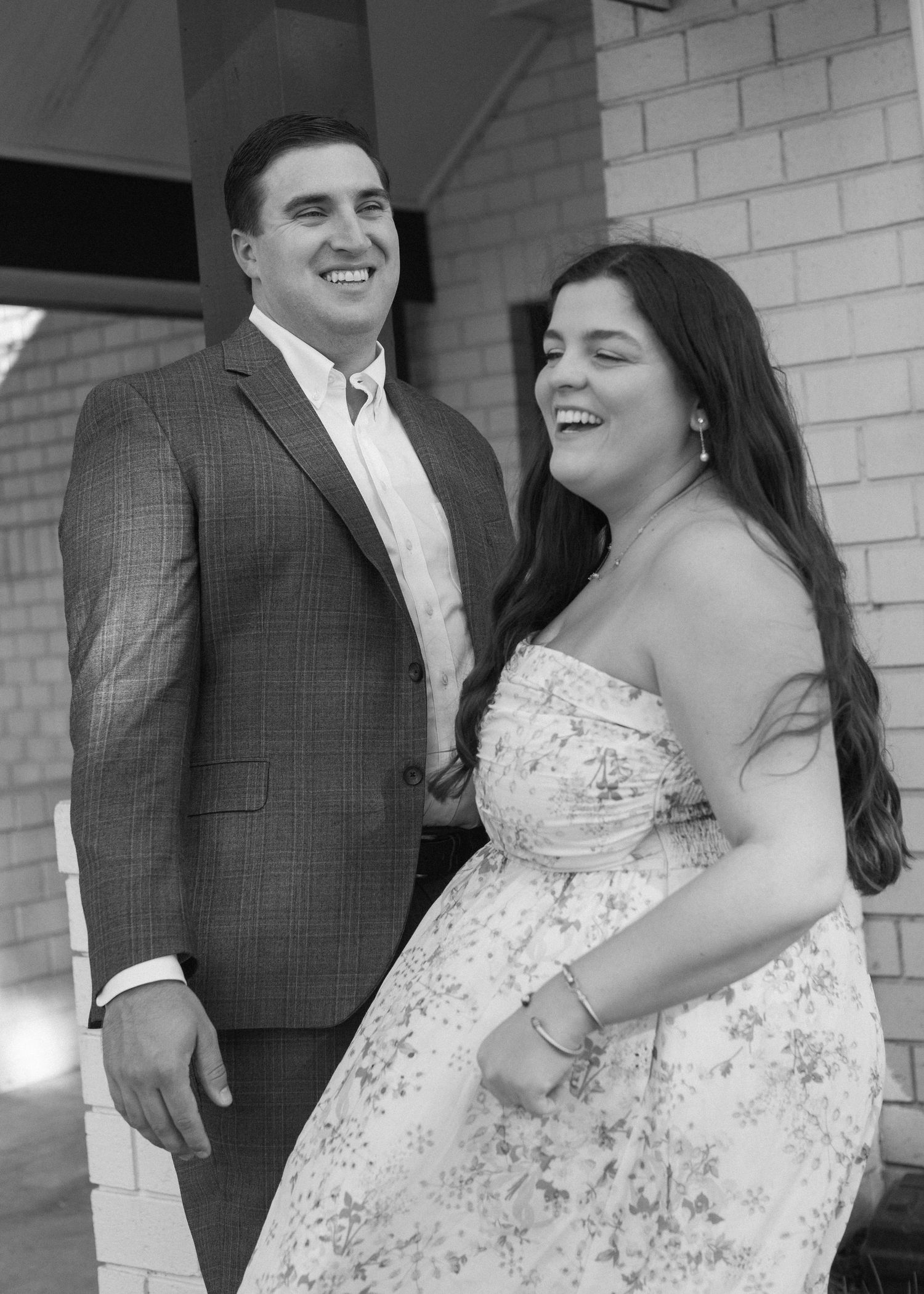 A couple shares a joyful moment together, with her in a white floral dress and him in a gray suit jacket.