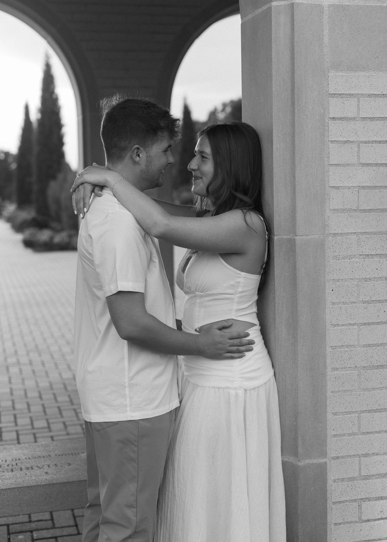A couple embraces under an archway with brick columns and a tiled walkway.