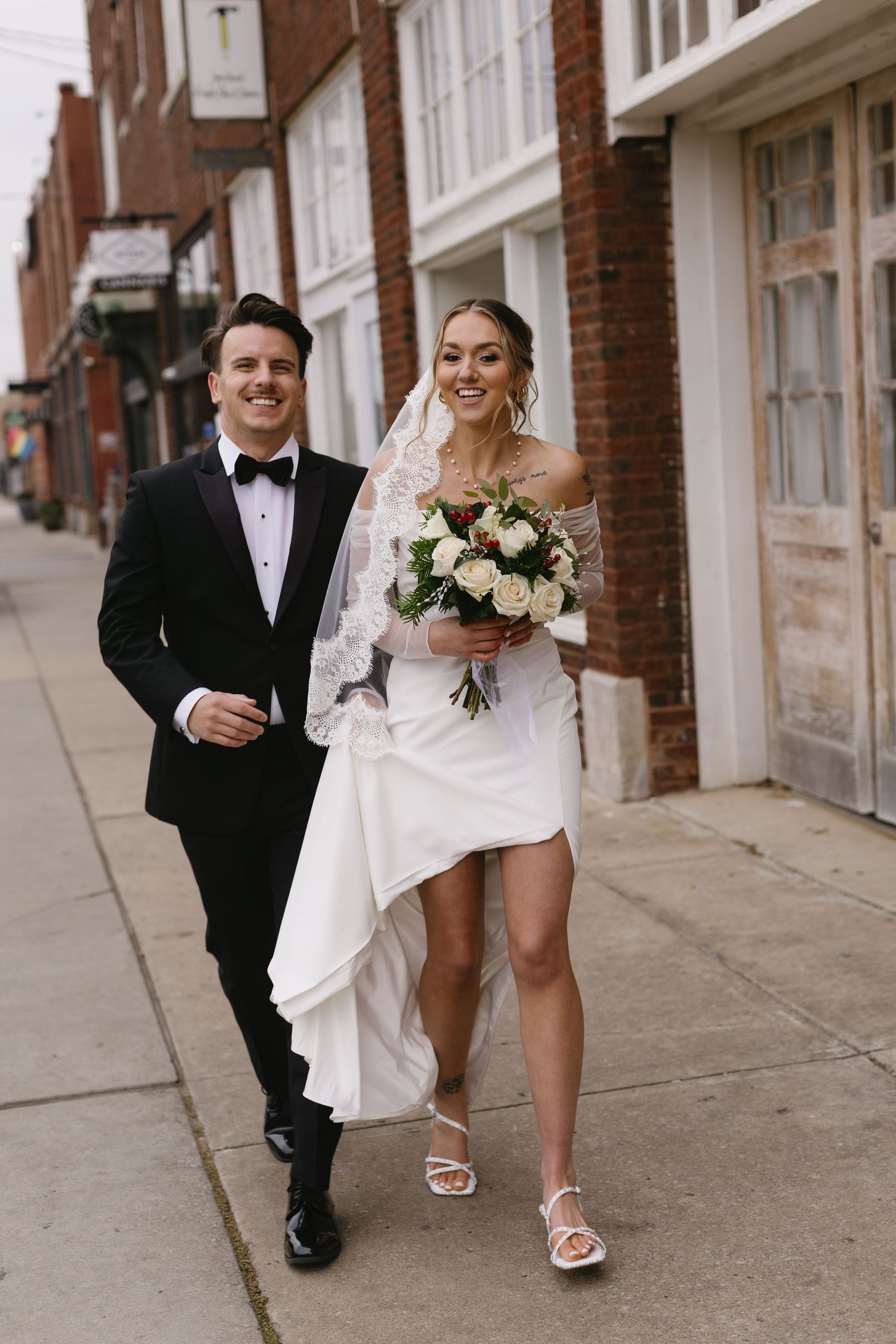 Newlyweds walking down city sidewalk with bride holding white bouquet.