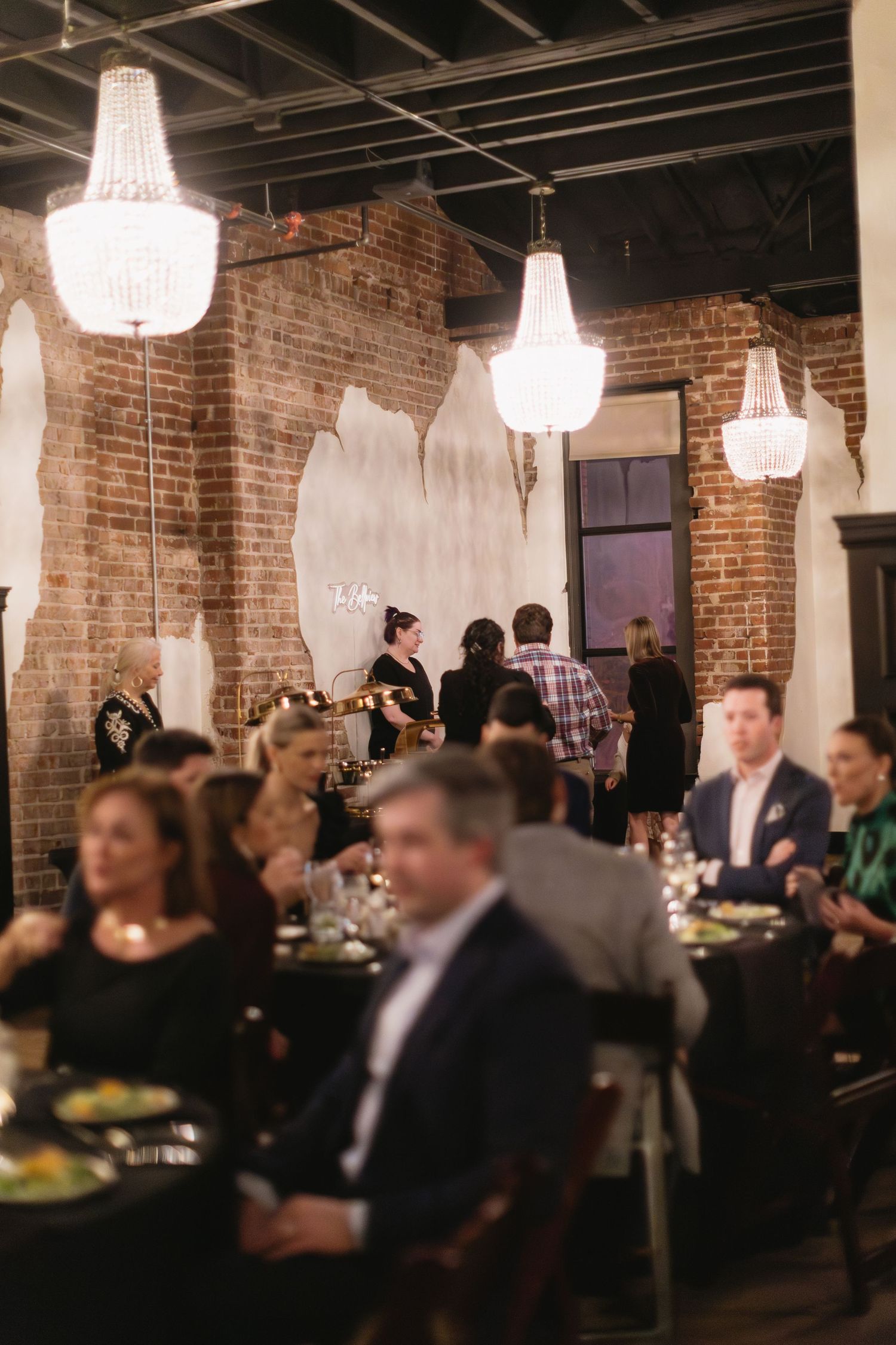 Large group celebrating in industrial-style venue with brick walls and crystal chandeliers.
