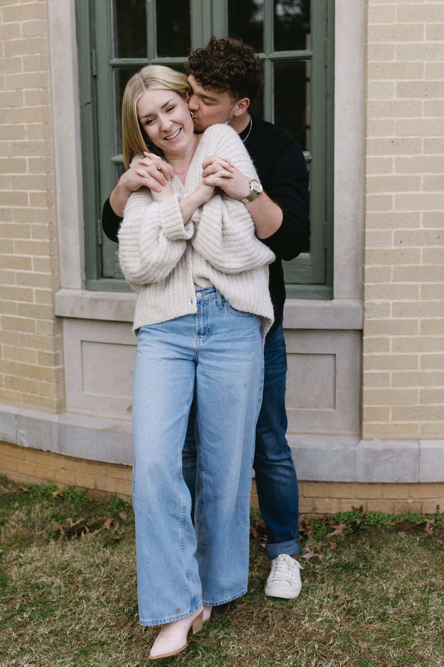 A tender moment shared between a couple in cozy sweaters and jeans against a brick building.